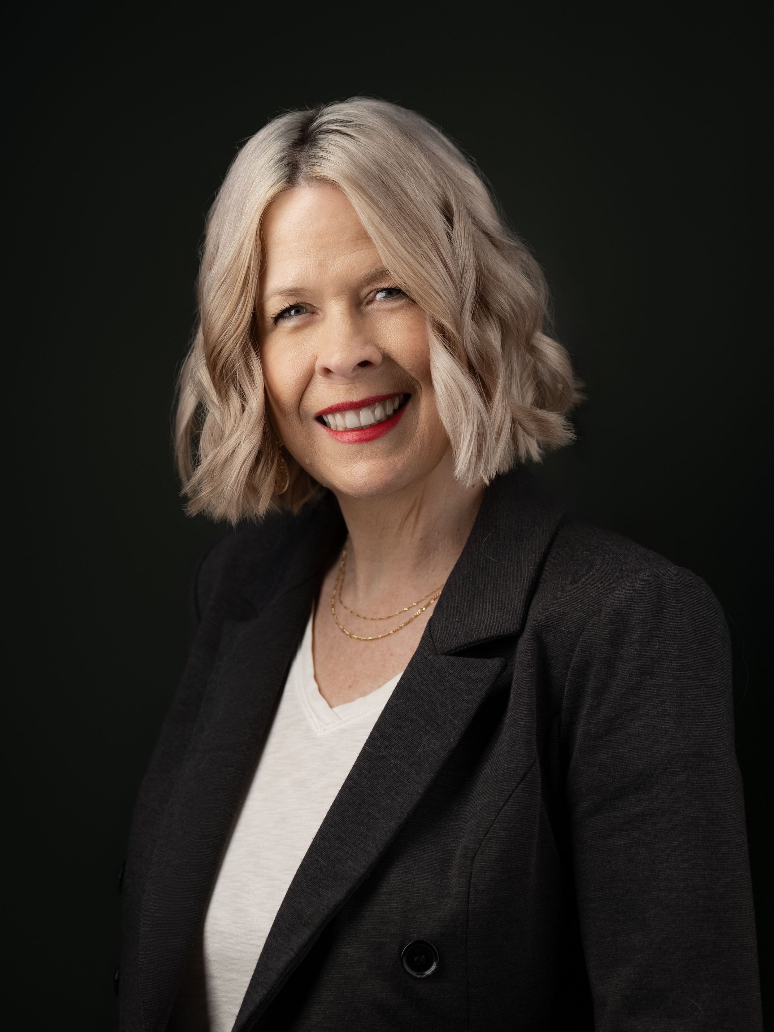 Professional portrait of a person with wavy silver-blonde hair wearing a black blazer and white top against dark background.