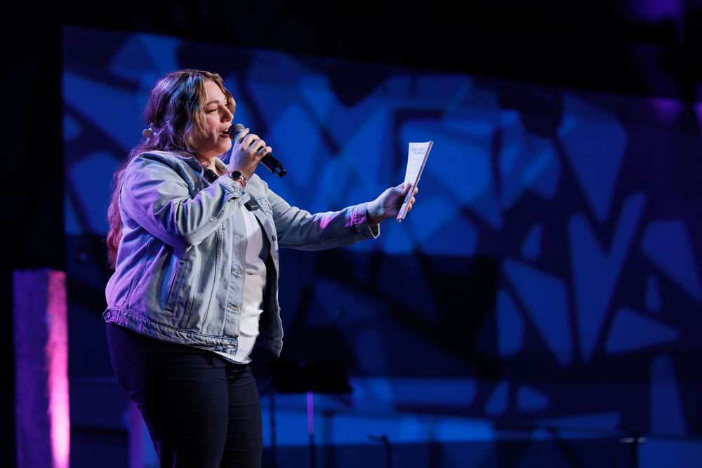 Presenter in jean jacket speaks into microphone while holding papers on illuminated stage.