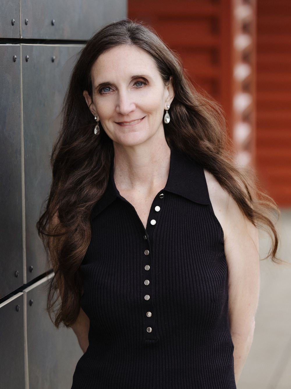 Professional headshot of a woman with long brown hair wearing a black sleeveless button-up top against metal and wood backdrop.