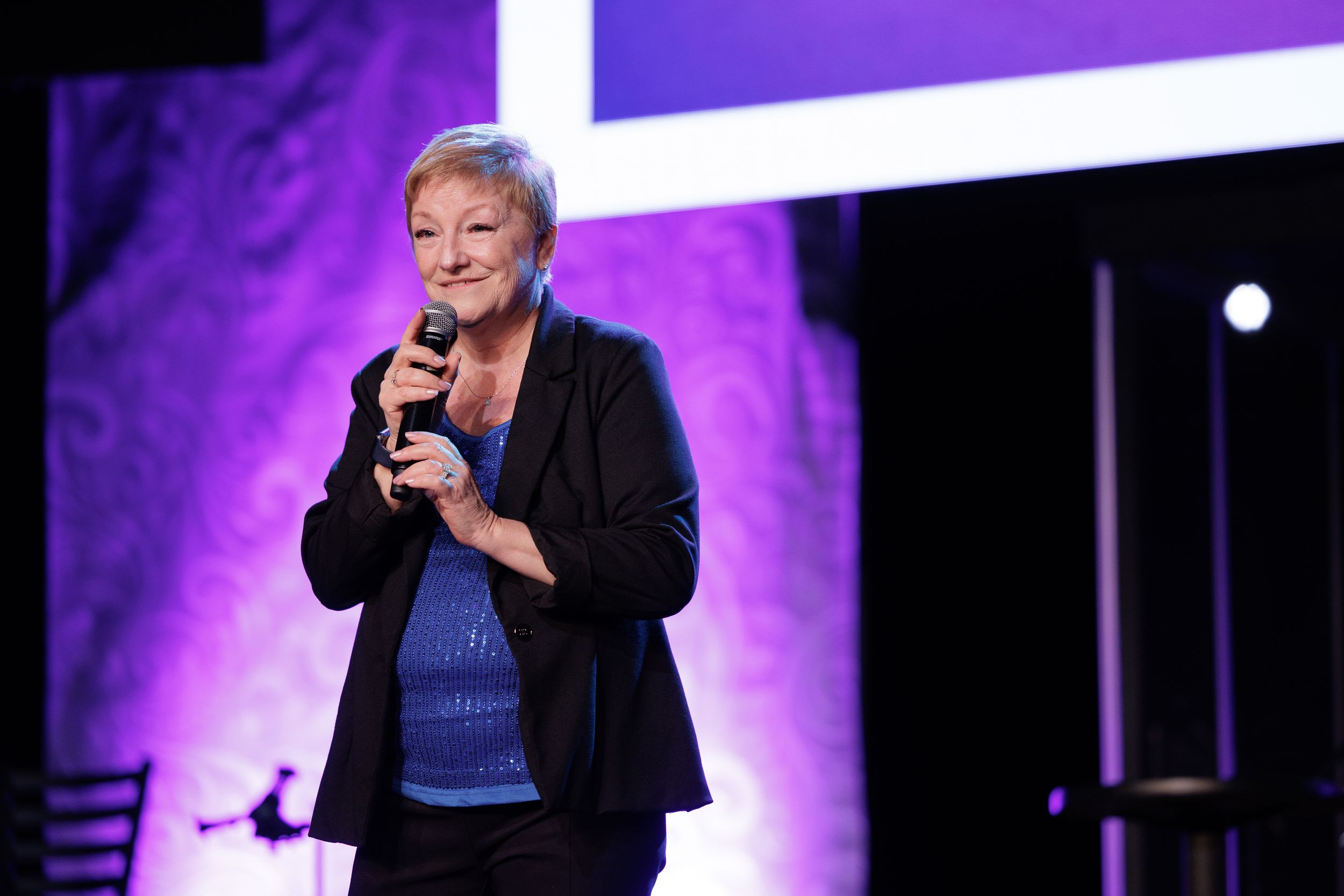 Performer on stage speaks into microphone with purple lighting and dark background during live show.