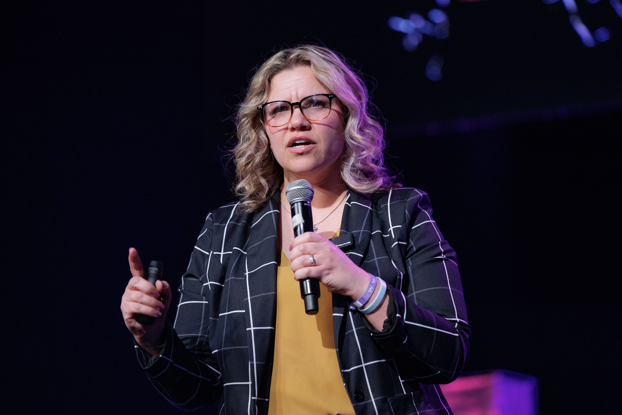 A speaker performs on stage wearing a yellow sweater and black plaid jacket while holding a microphone.