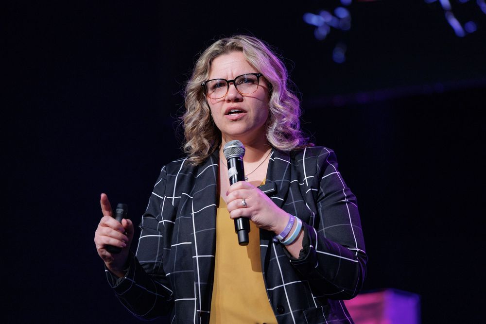 A speaker performs on stage wearing a yellow sweater and black plaid jacket while holding a microphone.