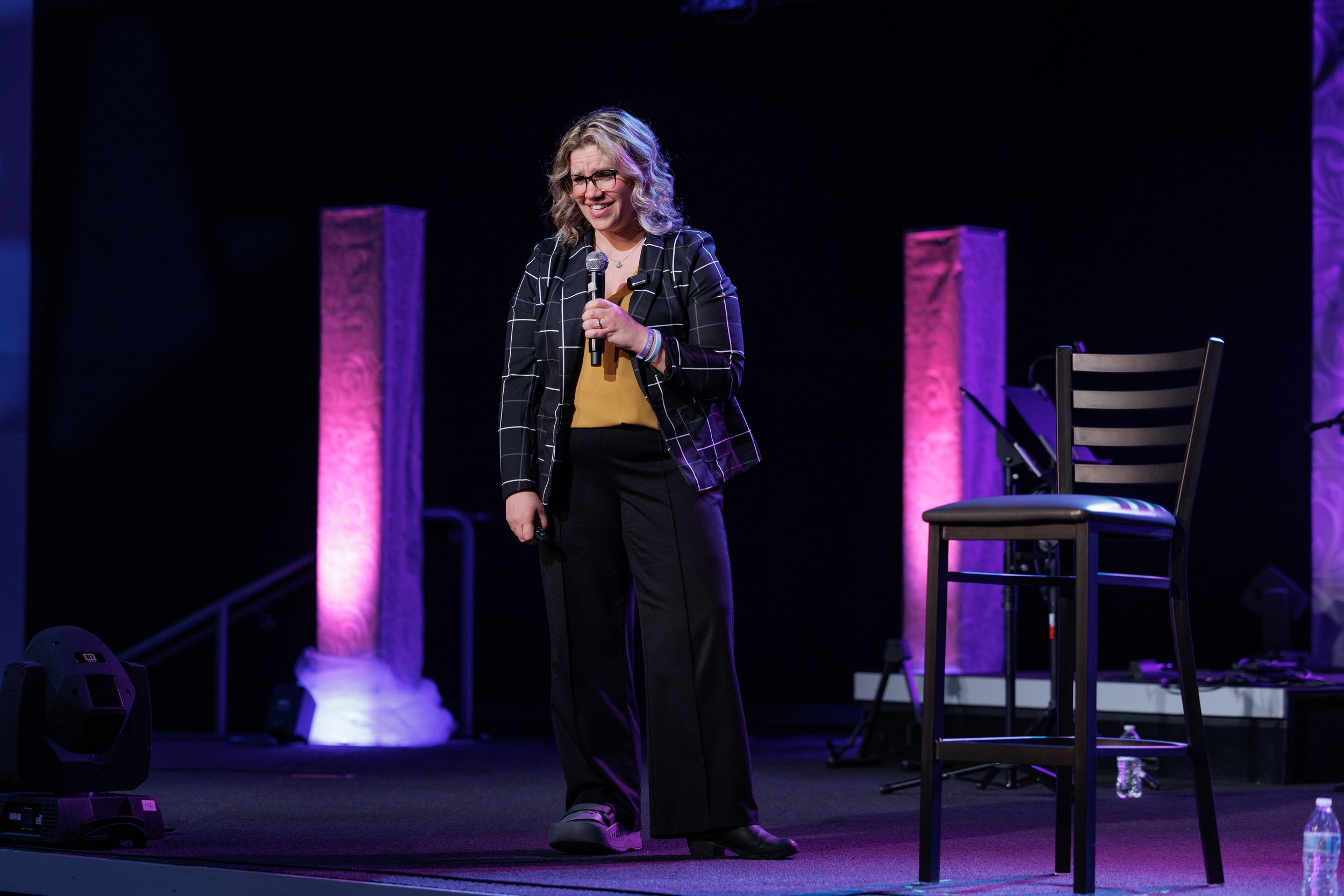 Speaker presents on stage with illuminated purple pillars and bar stool in dark theater setting.