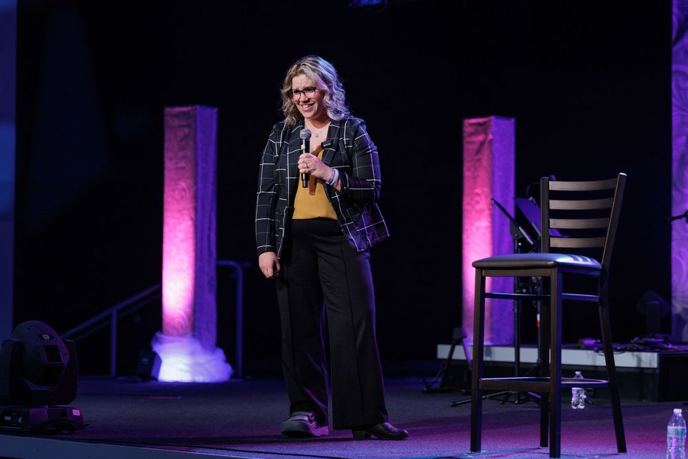 Speaker presents on stage with illuminated purple pillars and bar stool in dark theater setting.