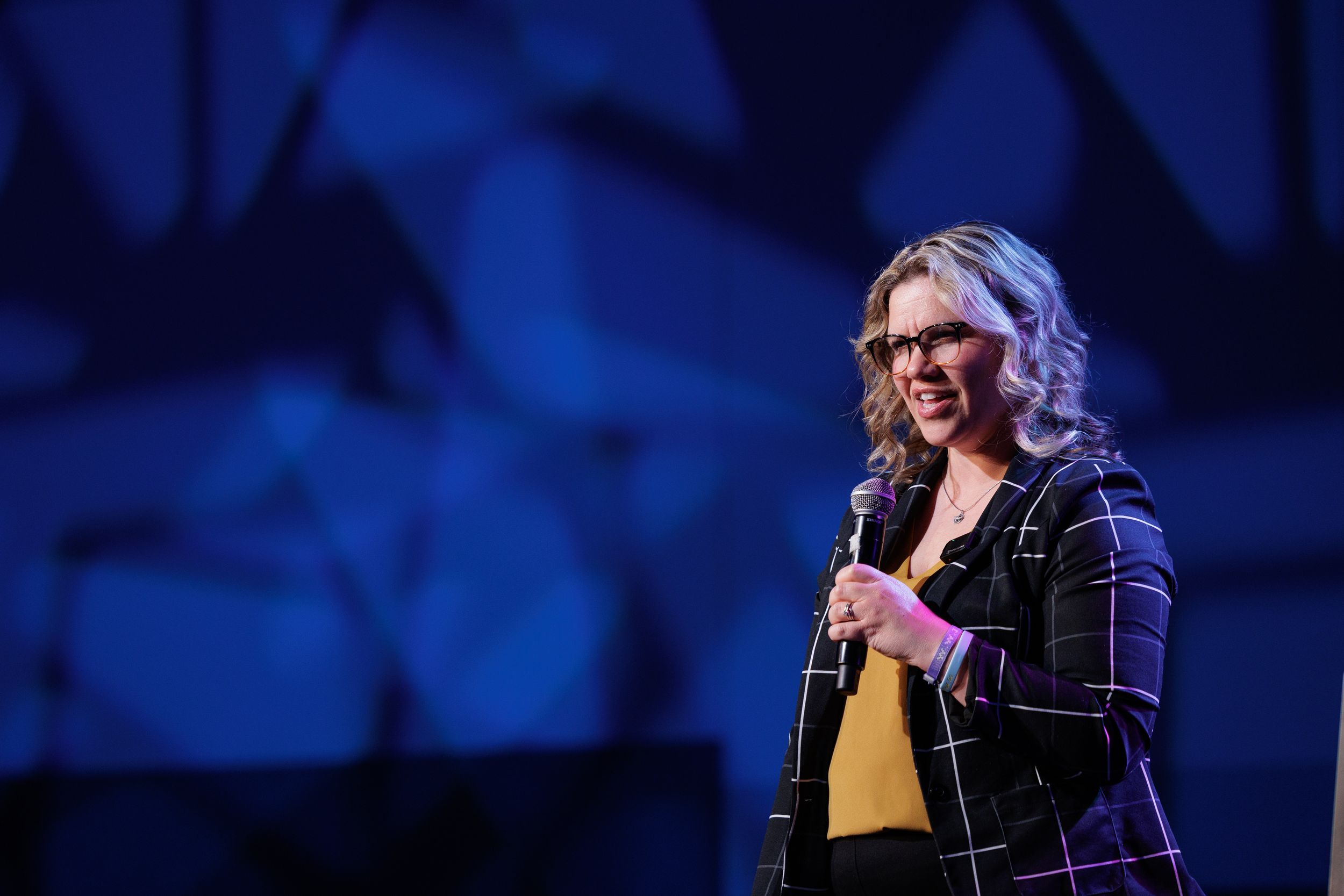 Speaker in plaid jacket performs on stage with dramatic blue lighting and geometric backdrop.
