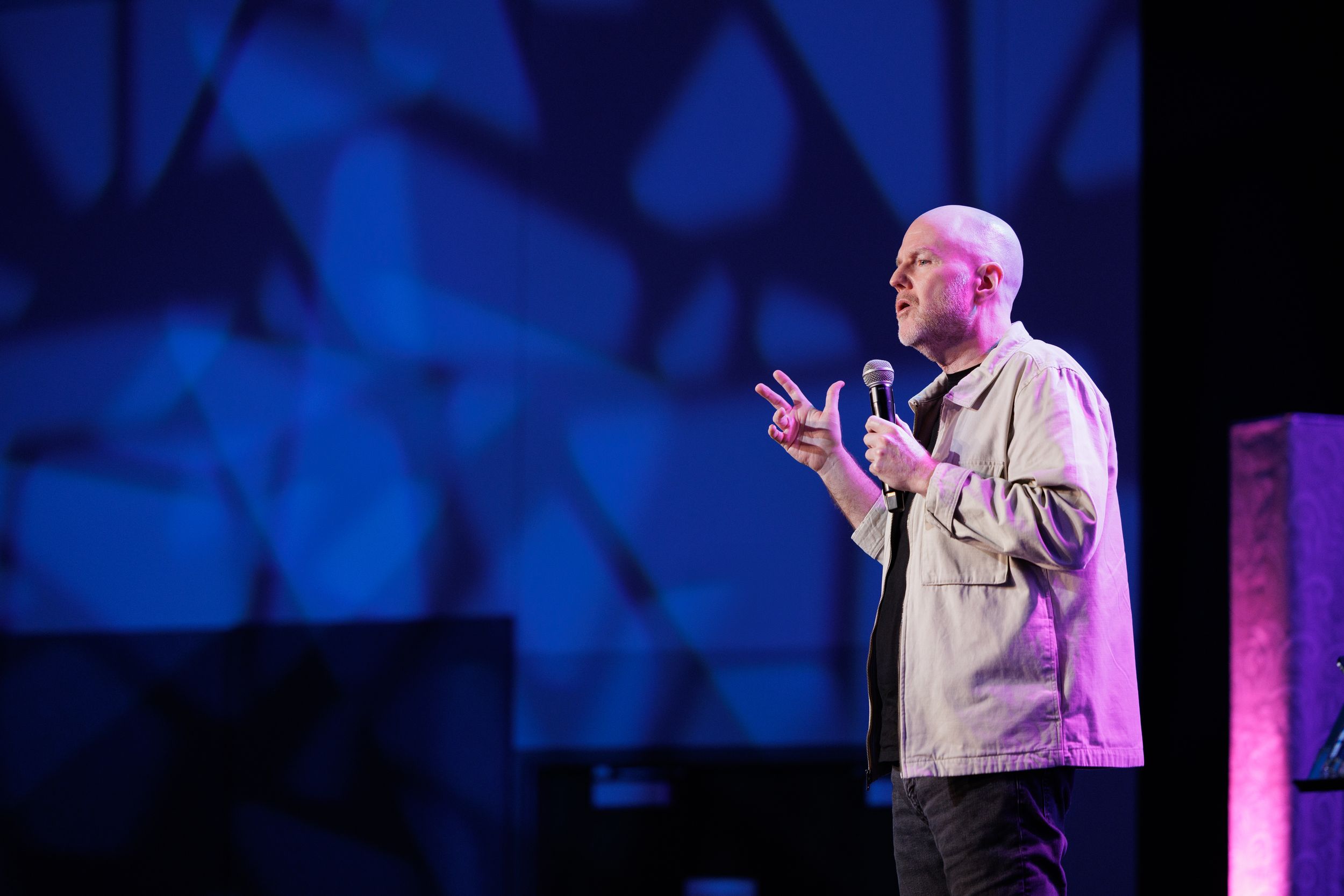 A speaker performs on stage with dramatic blue lighting and geometric patterns projected on the backdrop.
