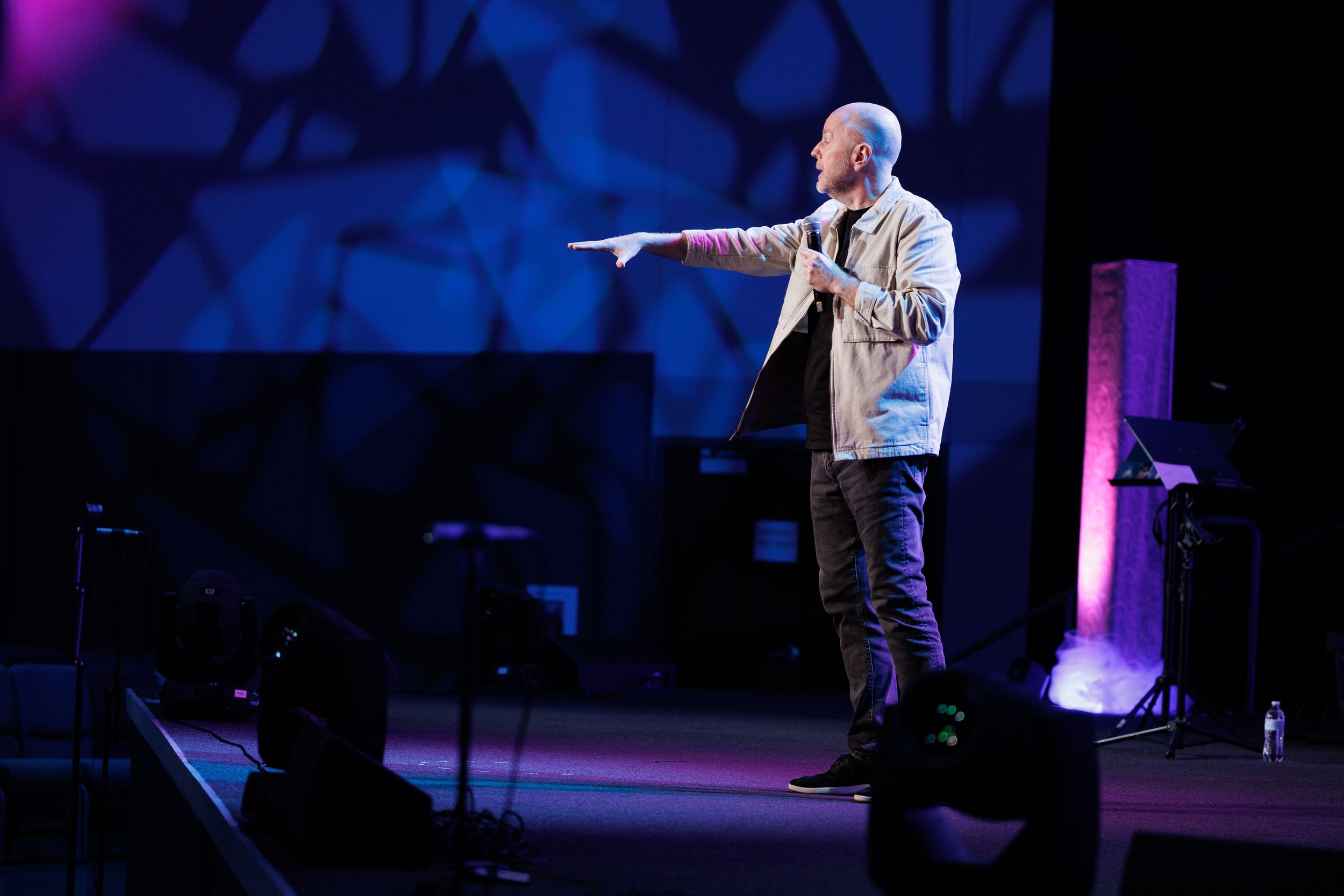 A performer in a light-colored jacket gestures on stage during a comedy show with blue lighting and geometric backdrop.