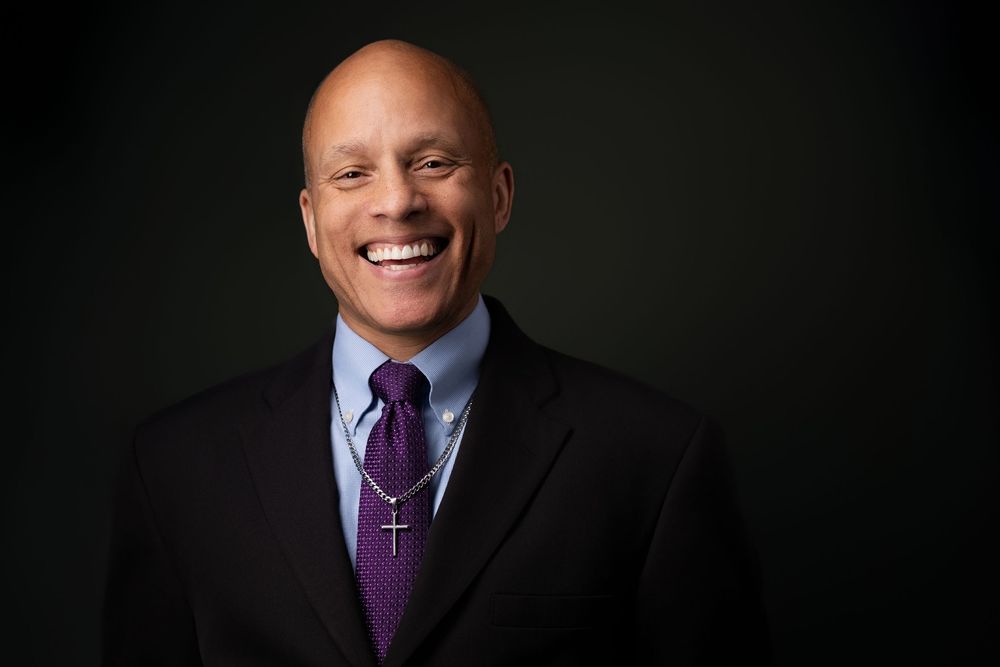 Professional headshot of person in dark suit and purple tie smiling against black background.