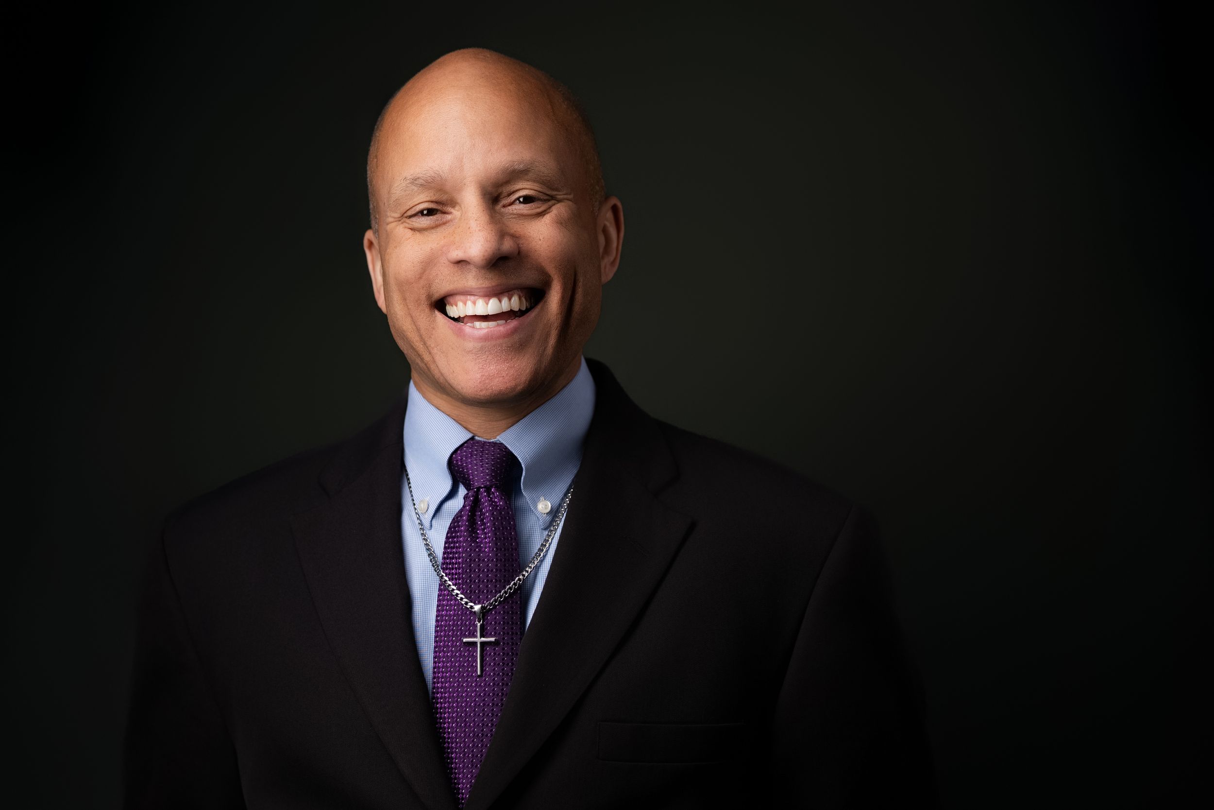 Professional headshot of person in dark suit and purple tie smiling against black background.