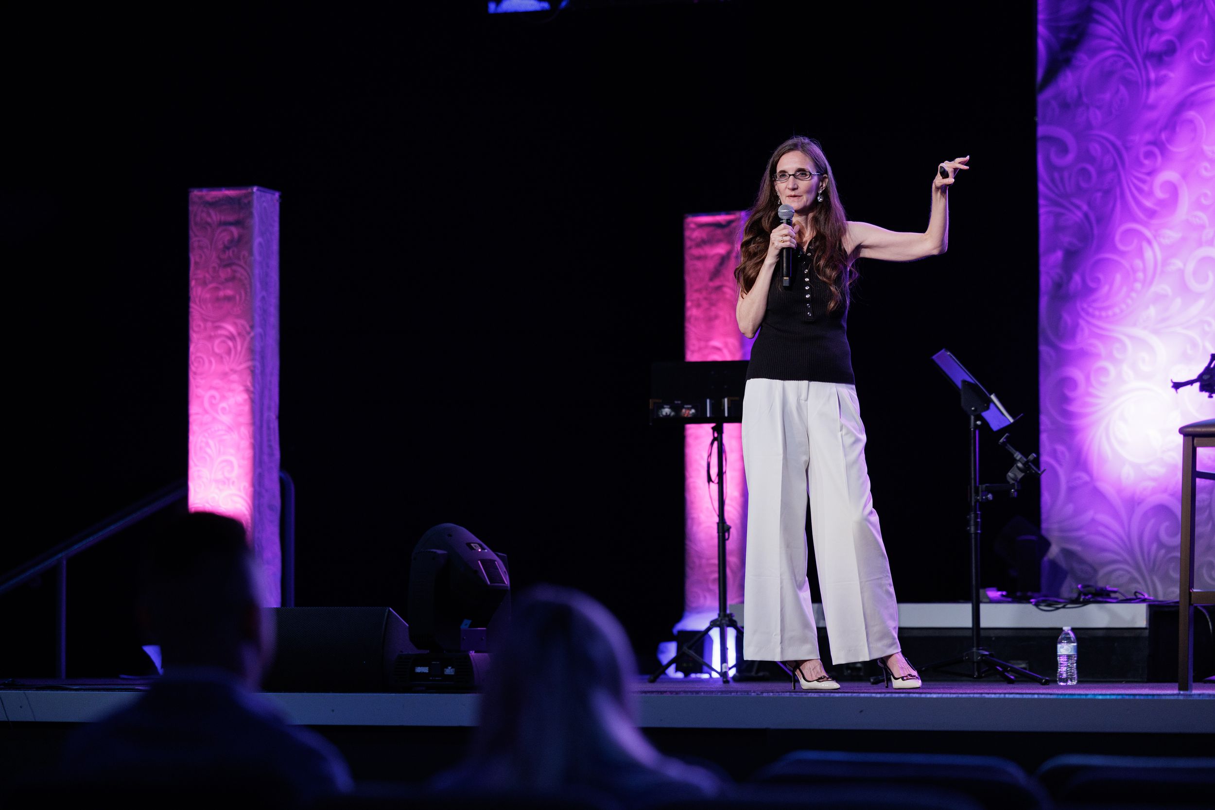 A performer in black top and white pants stands on a dimly lit stage with purple lighting effects.