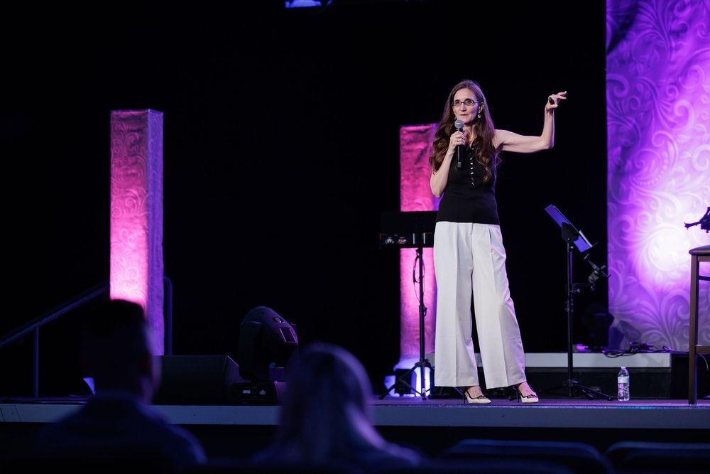 A performer in black top and white pants stands on a dimly lit stage with purple lighting effects.