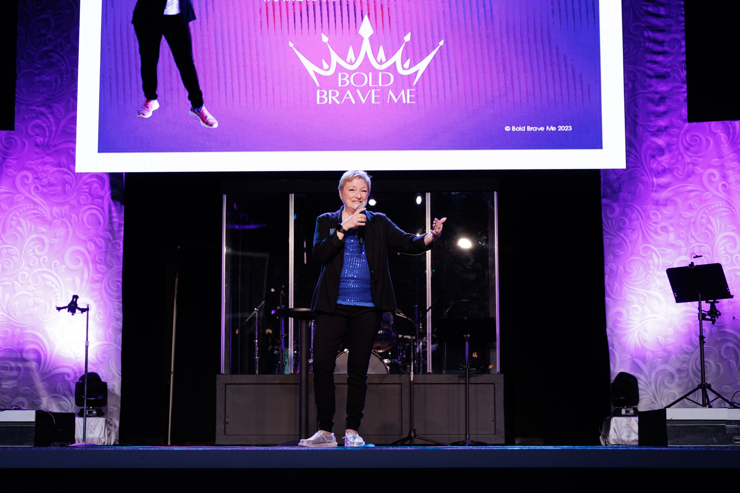 A speaker stands on stage with dramatic purple lighting and a large screen display behind them.