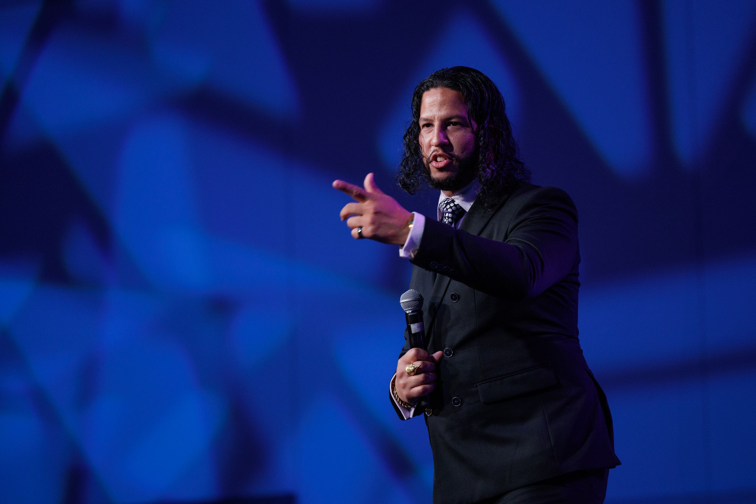 Performer in dark suit gestures while speaking on stage with blue geometric backdrop.