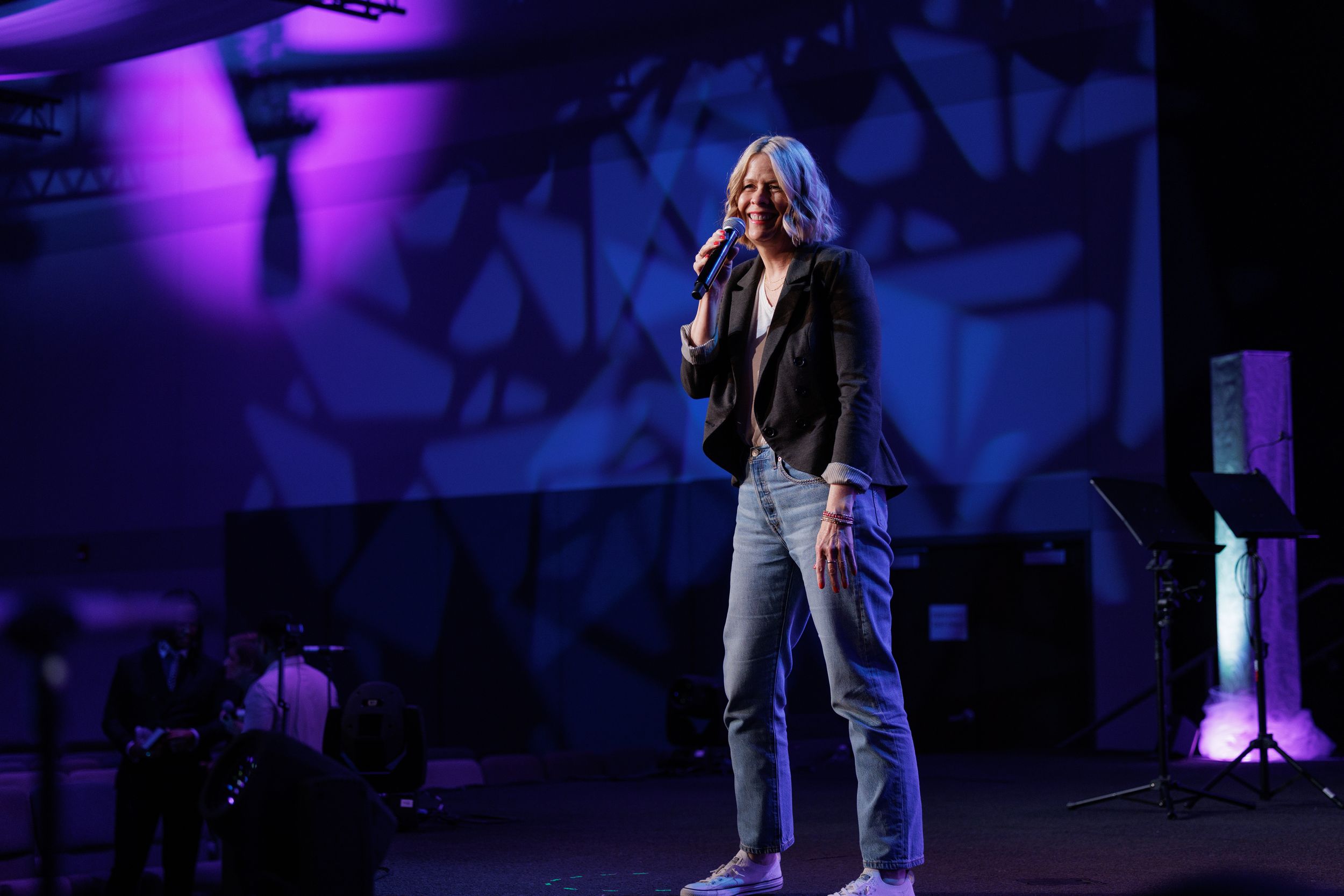 A performer in a leather jacket and jeans sings on stage with dramatic purple and blue lighting.