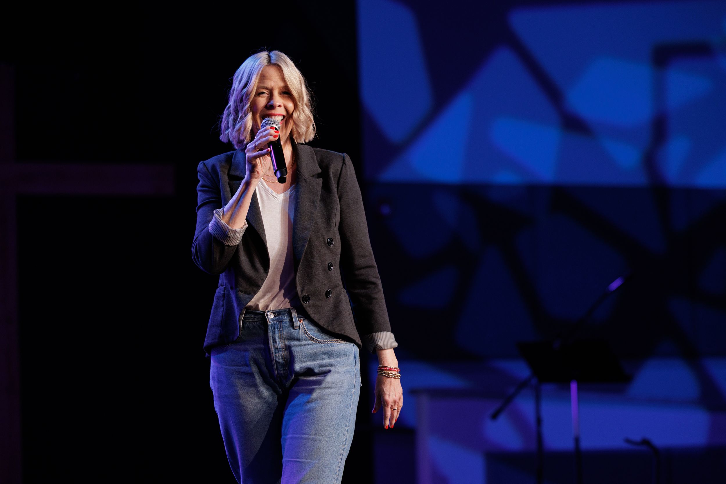 A performer wearing a blazer and jeans sings on stage with blue lighting in the background.