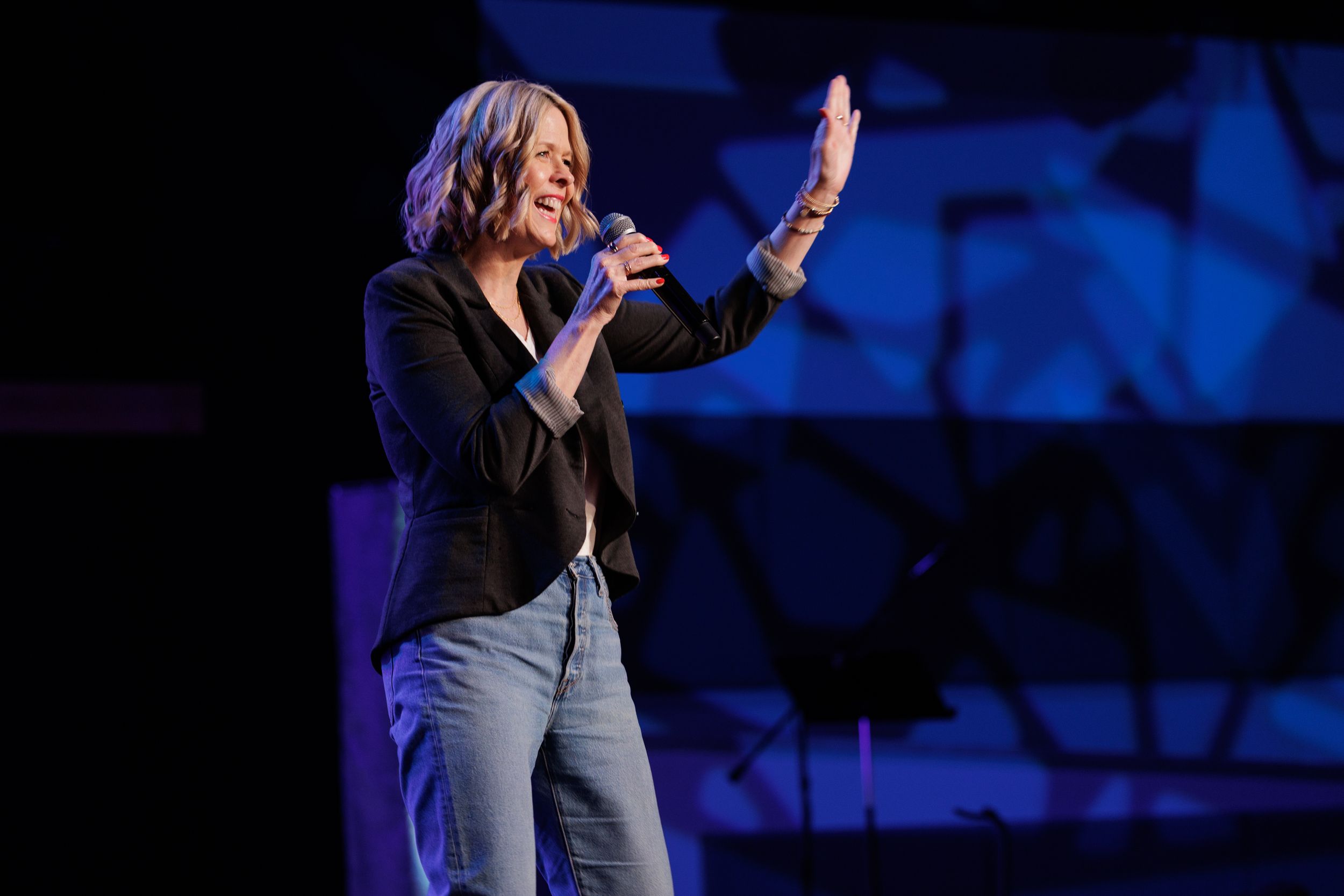 A speaker performs on stage in jeans and a black jacket against a blue-lit backdrop at a live event.