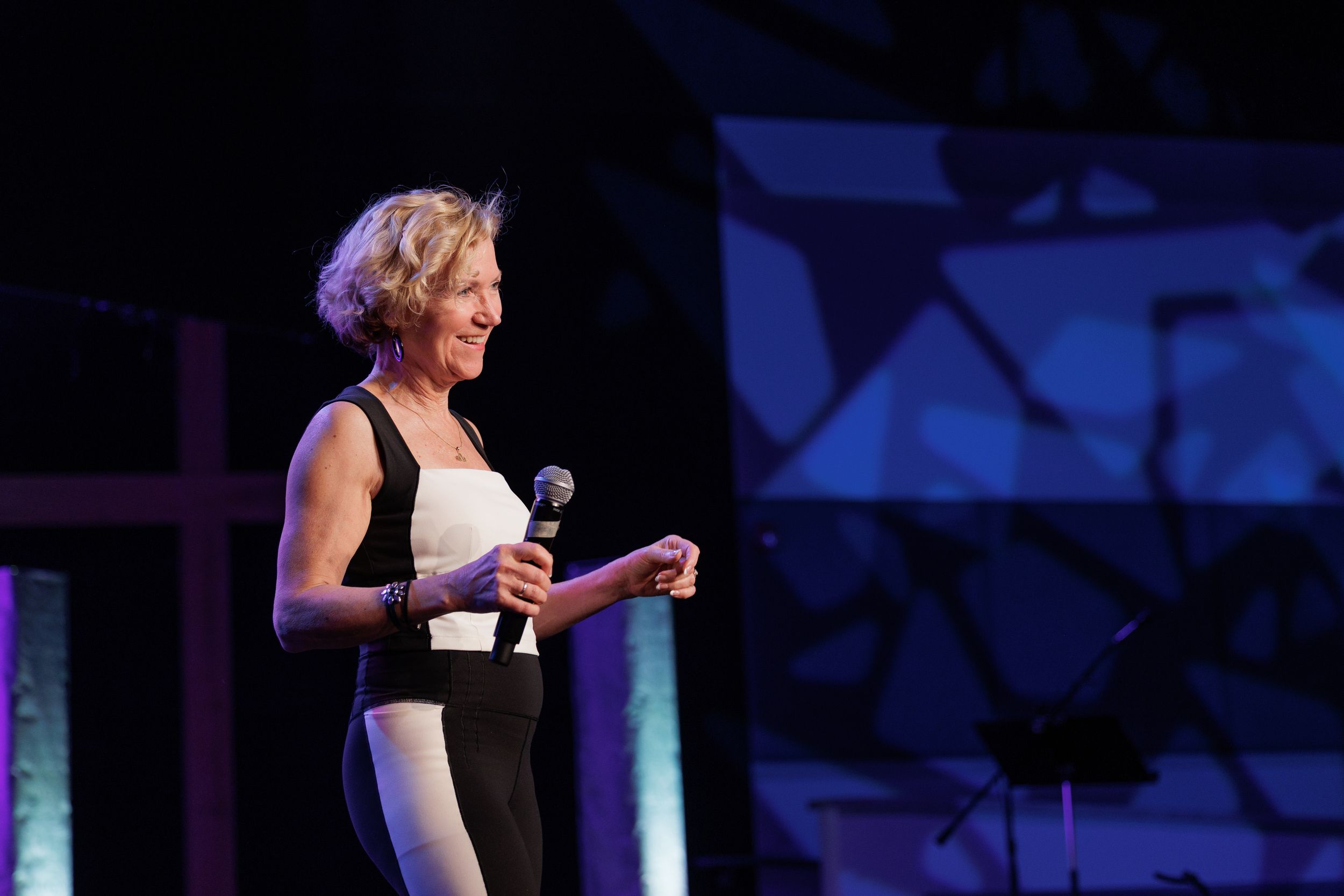 Presenter in black and white outfit speaks into microphone on stage with geometric patterns behind.