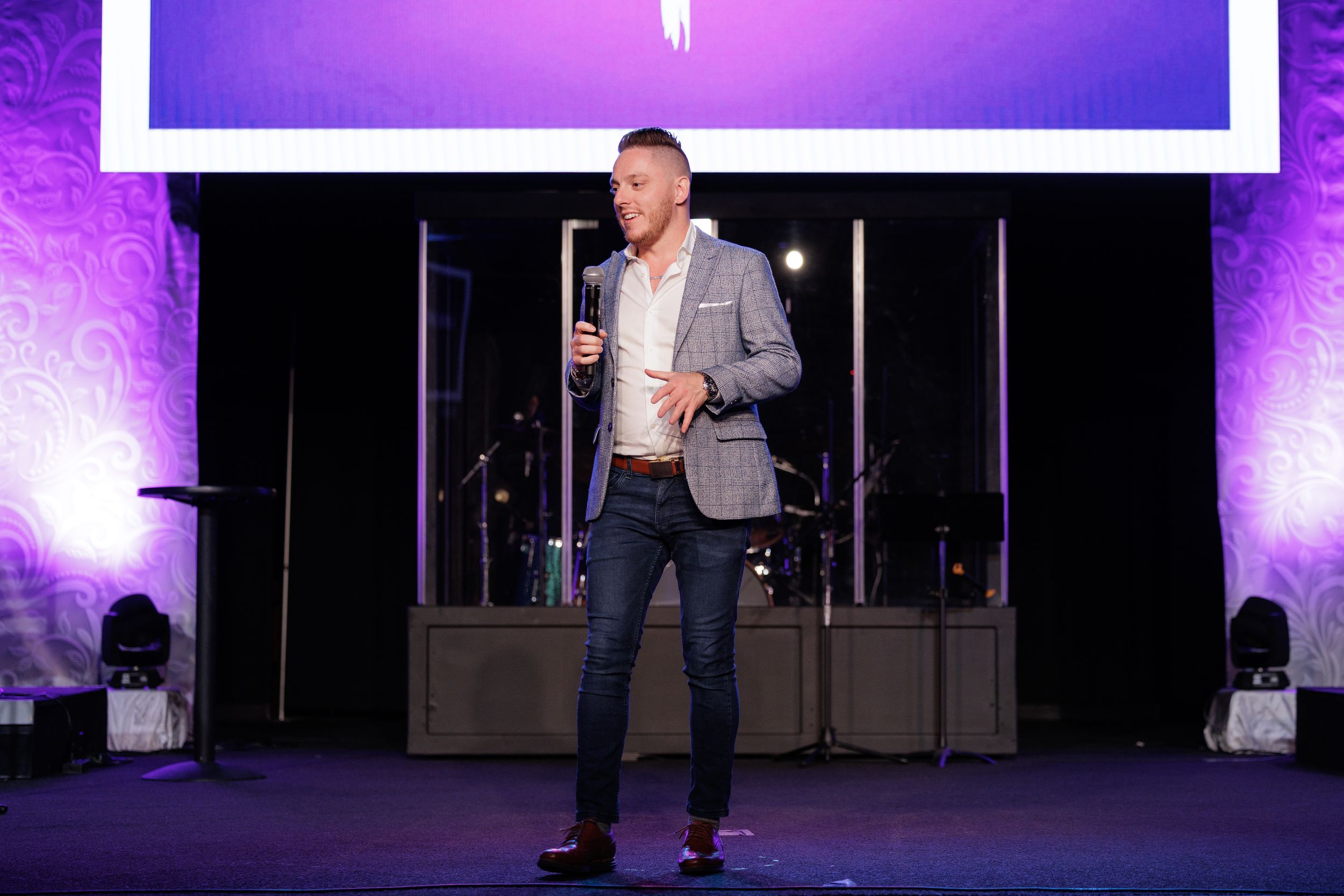Speaker in grey blazer presents on stage with purple-lit backdrop and audio equipment visible.