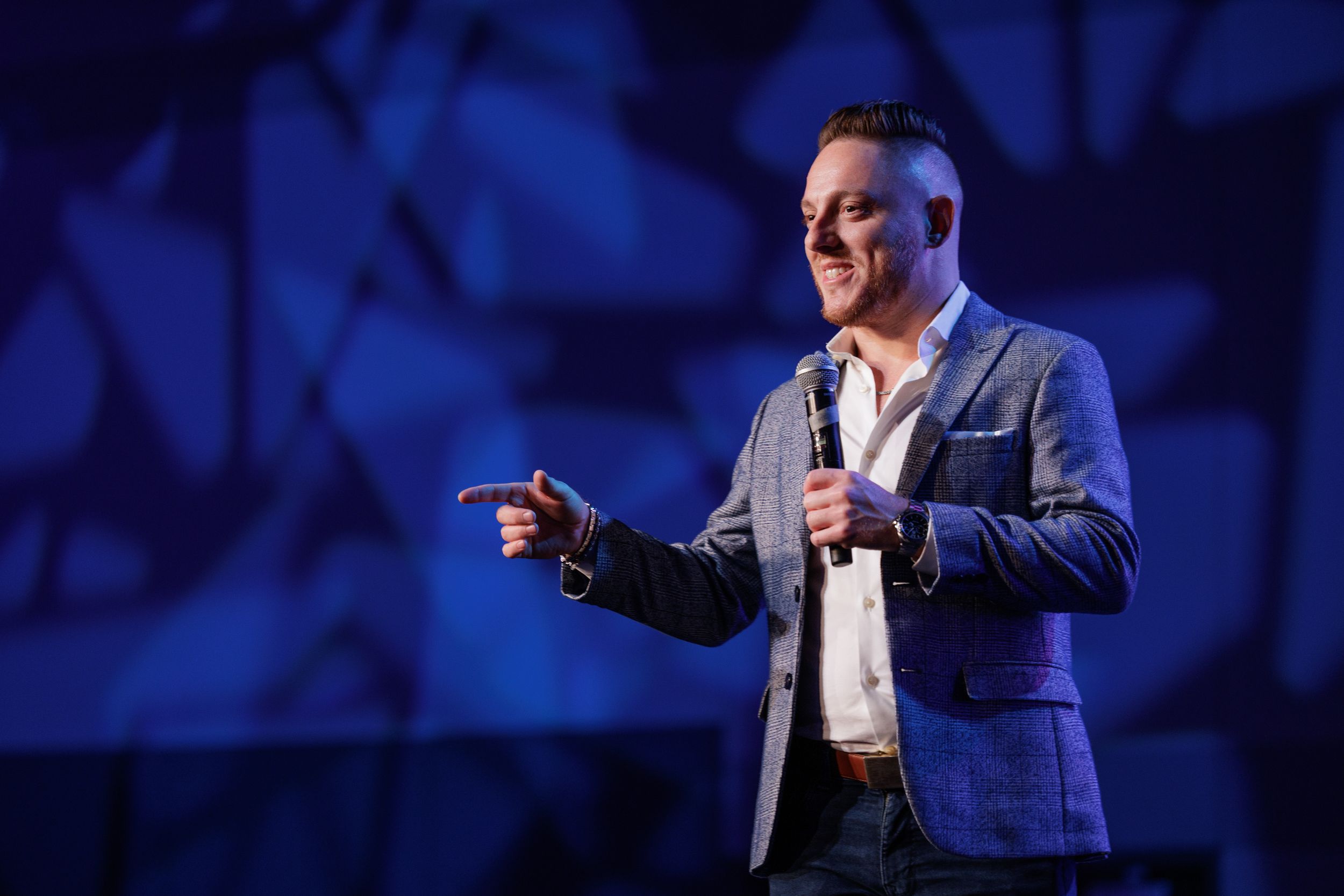 Speaker on stage wearing a blue blazer and white shirt gestures during presentation with blue lighting background.