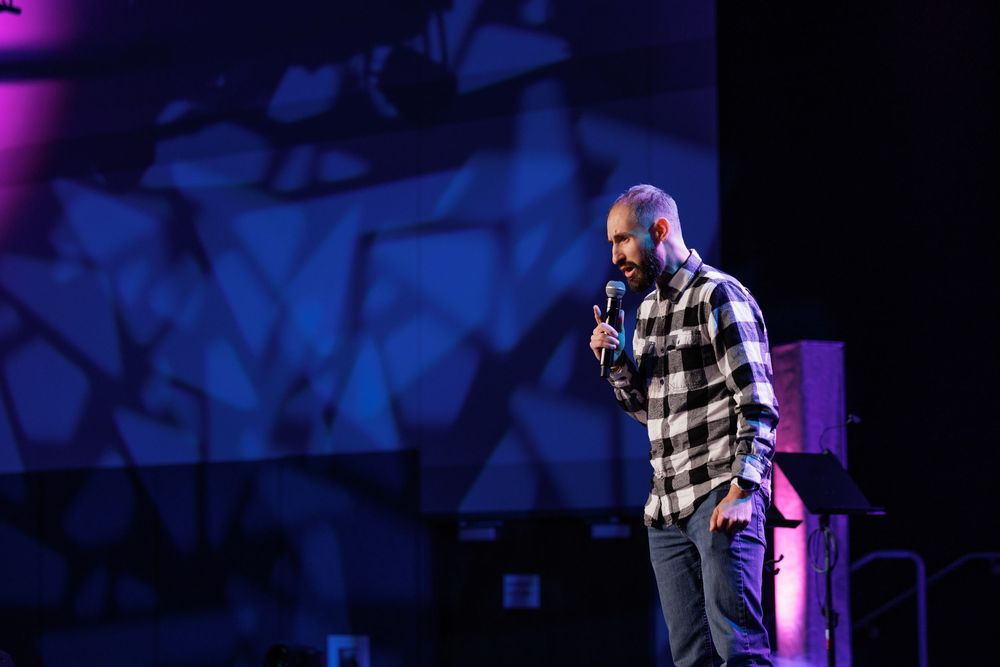 A speaker in a plaid shirt performs on stage with purple lighting and shadow effects.