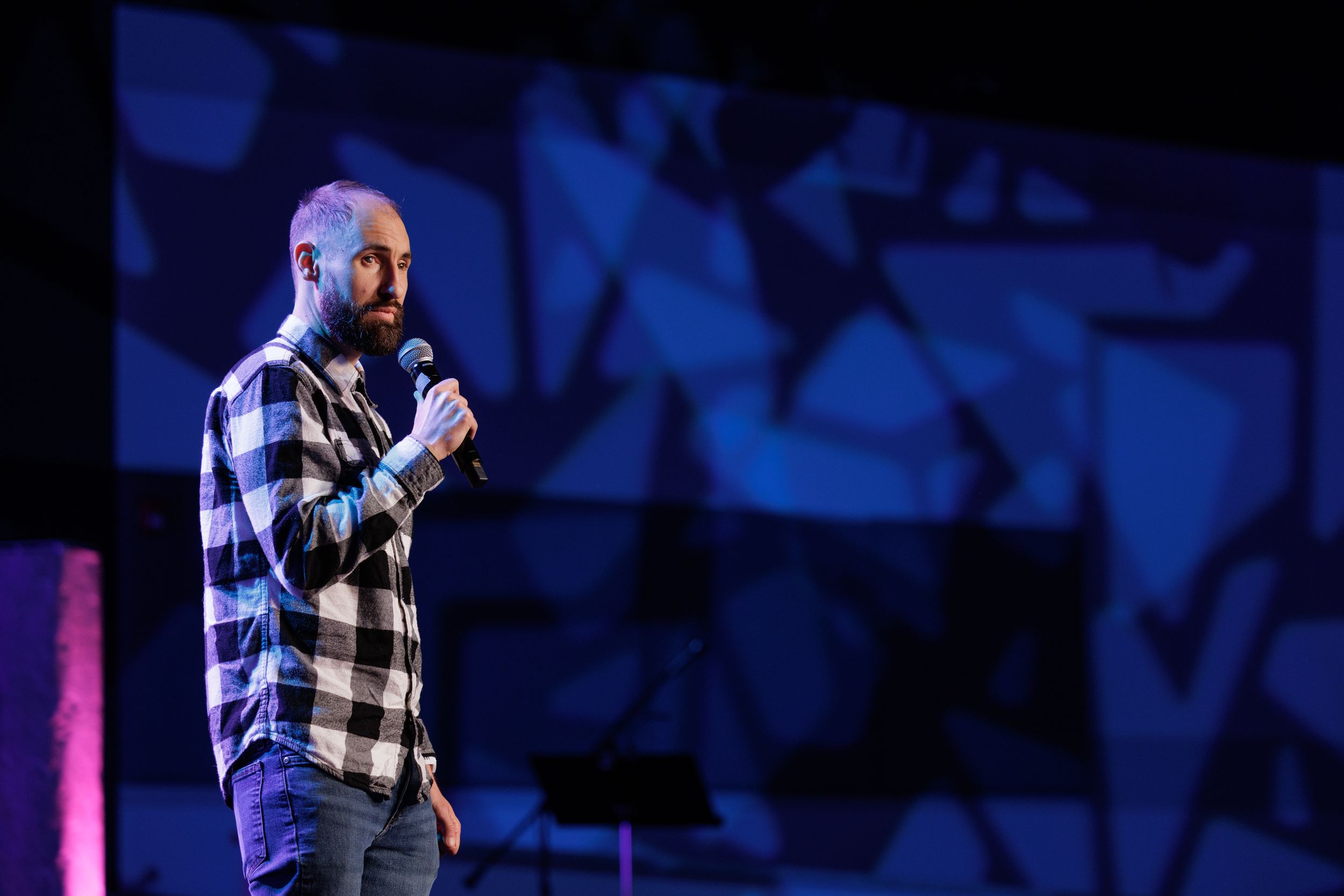 Performer in a blue and white plaid flannel shirt speaks into microphone on stage with blue geometric backdrop.
