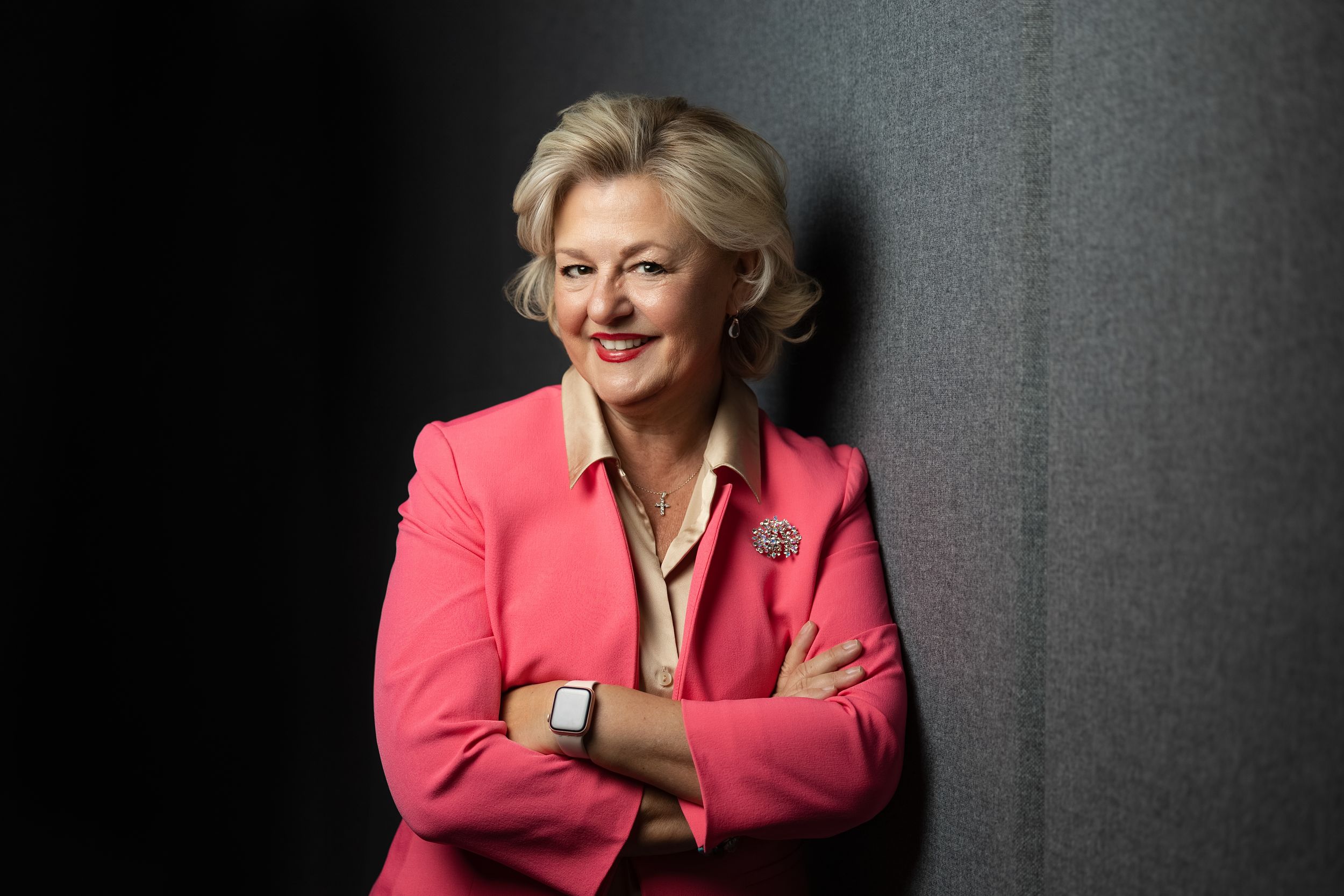 Business professional in a coral pink blazer poses against a gray background with dramatic side lighting.