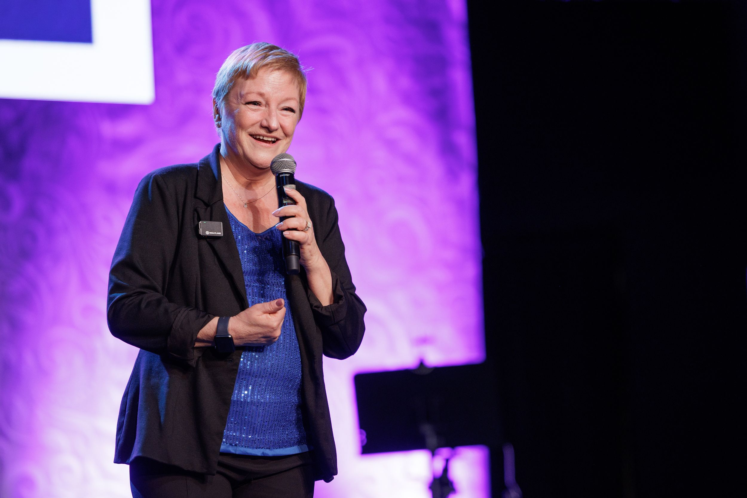 A presenter speaks into a microphone on stage with purple lighting in the background.