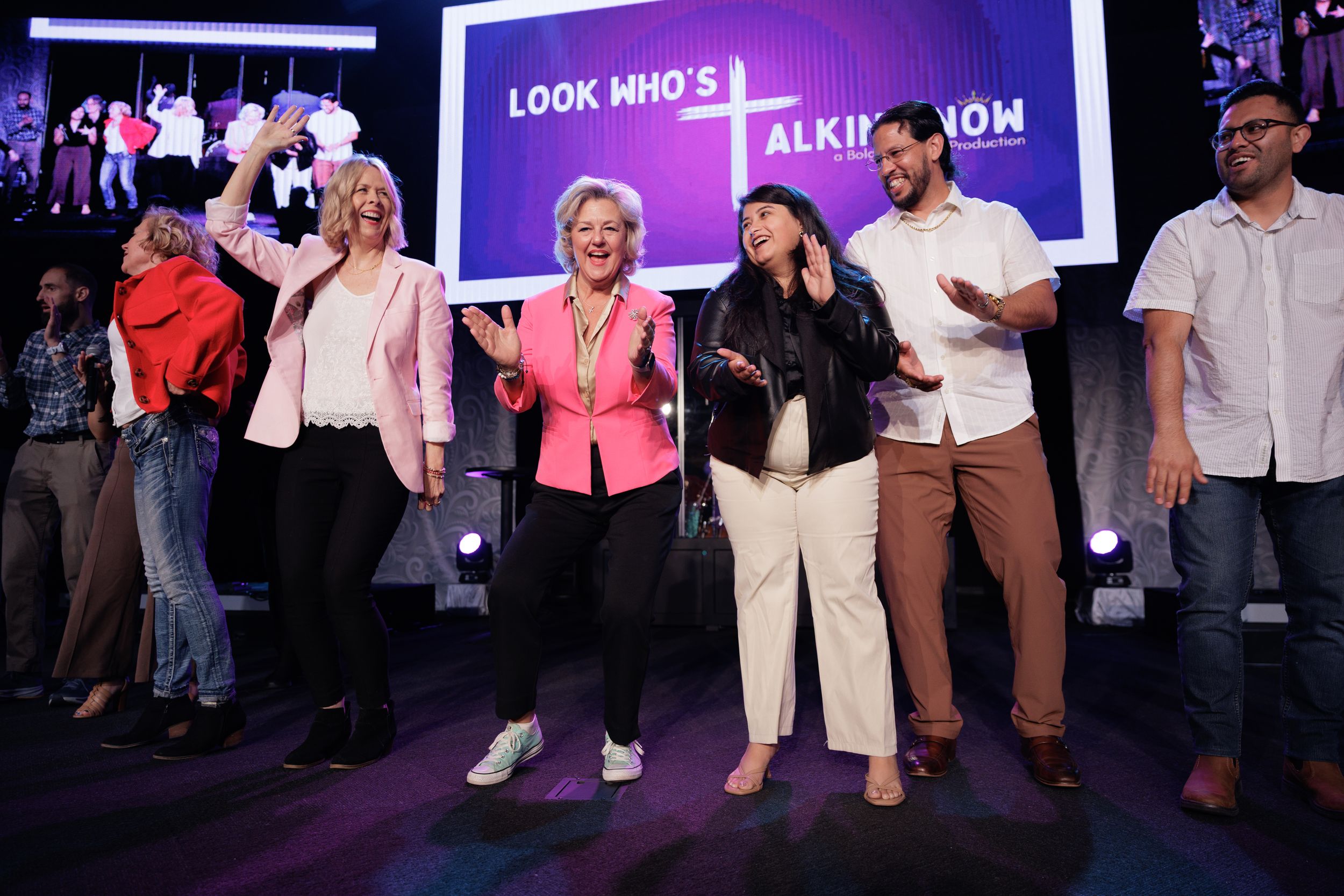 People on stage engage energetically with a purple-lit backdrop displaying text during a lively event presentation.