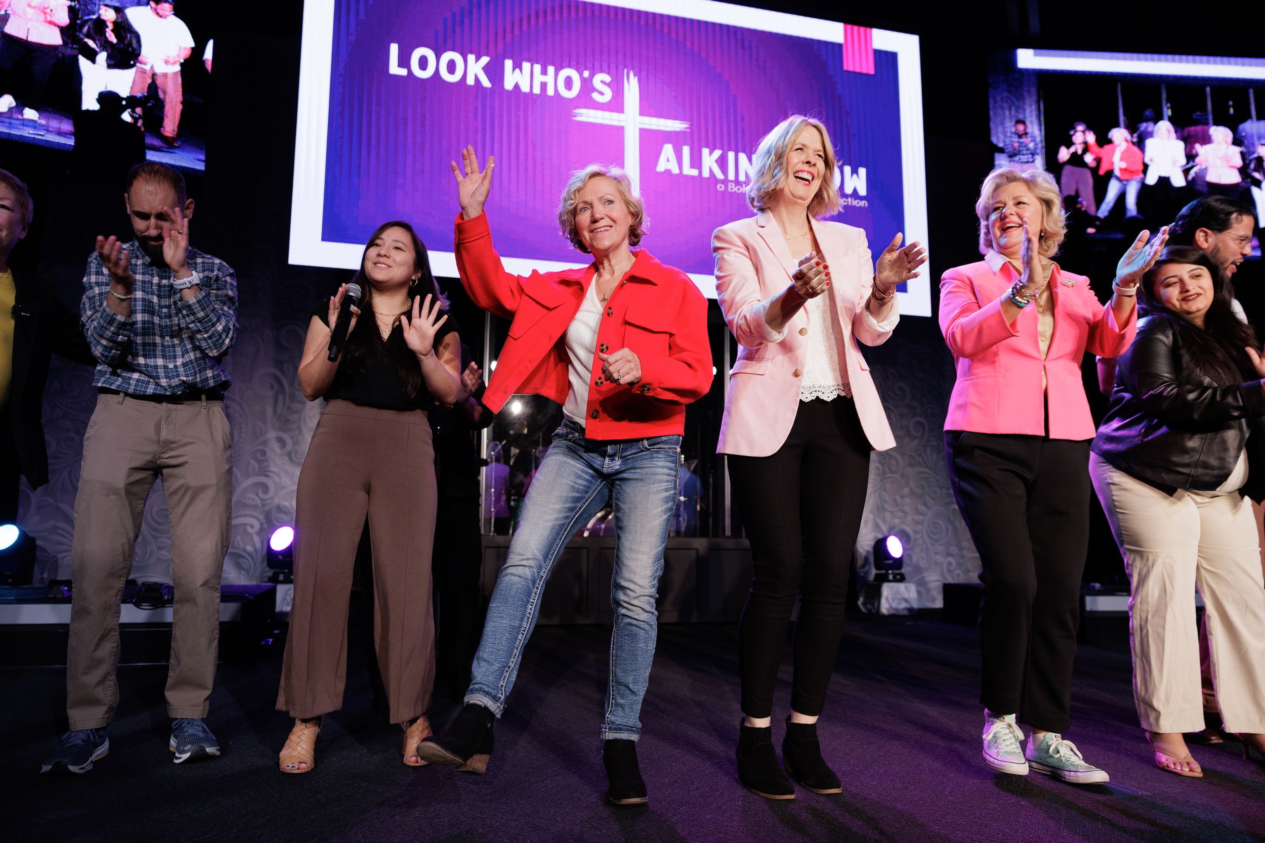 Group performs enthusiastically on stage with purple lighting and digital display screen in background during live event.
