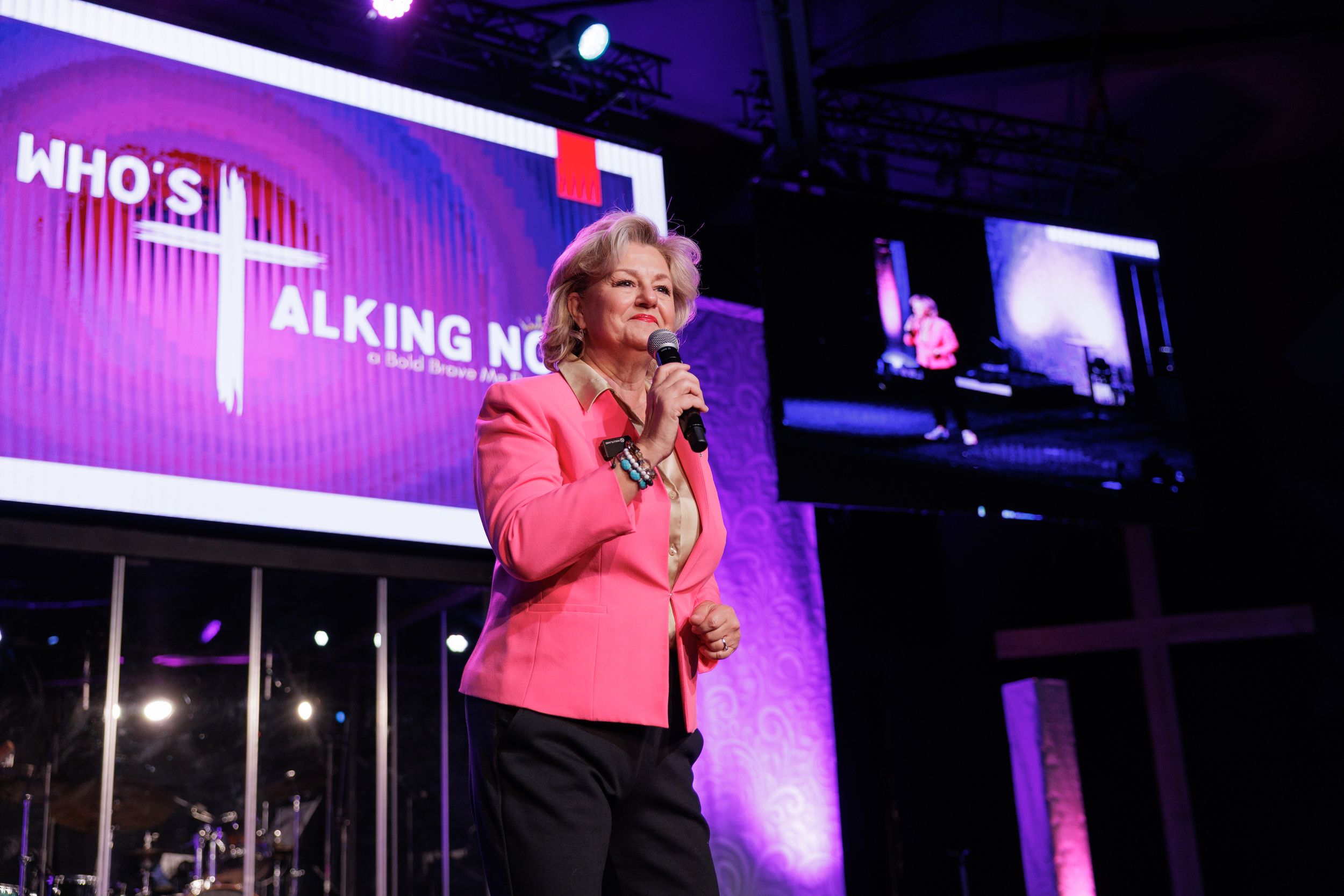 Professional speaker in pink blazer presents on stage with colorful backdrop and event signage.