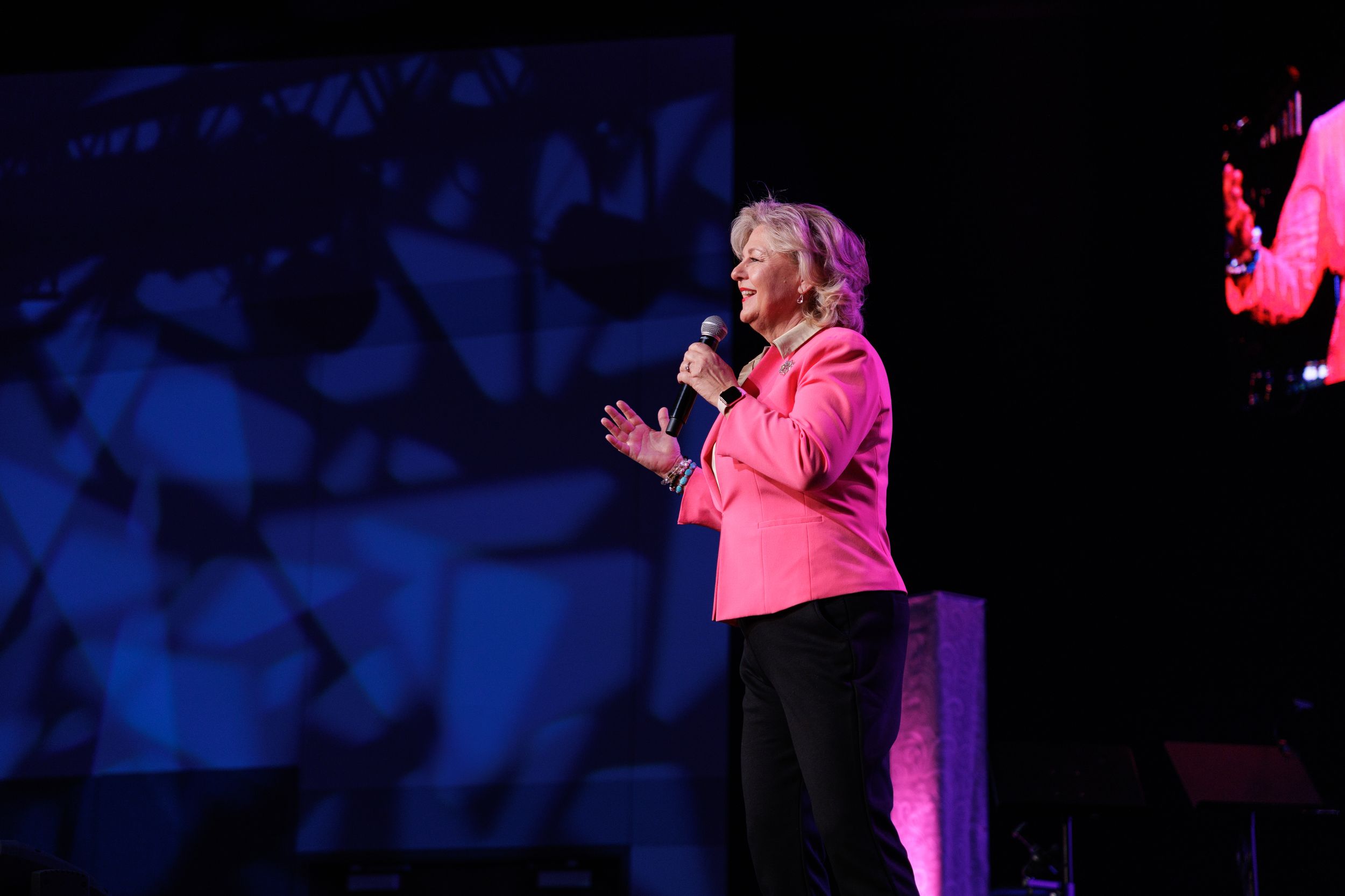 A speaker in a bright pink blazer addresses an audience from a dark stage illuminated by blue and pink lighting effects.