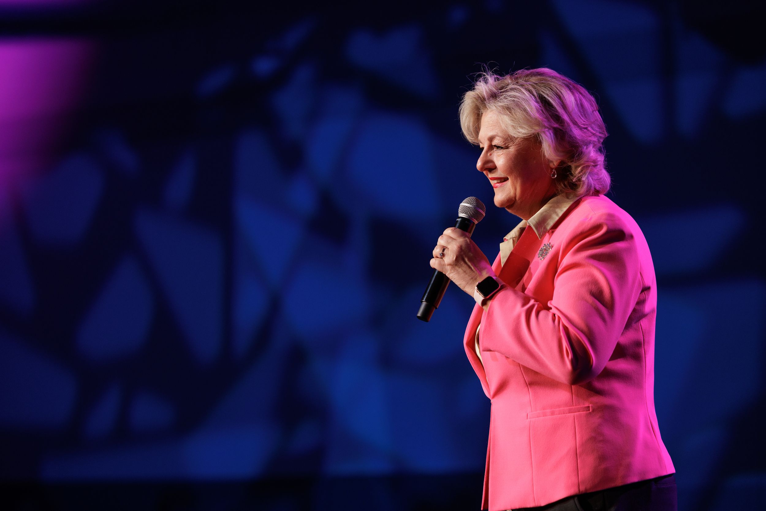 Speaker in bright pink blazer holds microphone while presenting on stage with blue lighting effects in background.