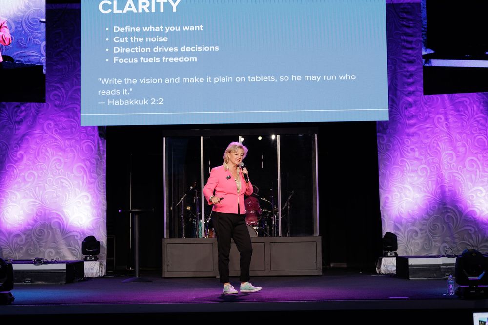 A presenter speaks on stage with a presentation slide about clarity displayed behind them.