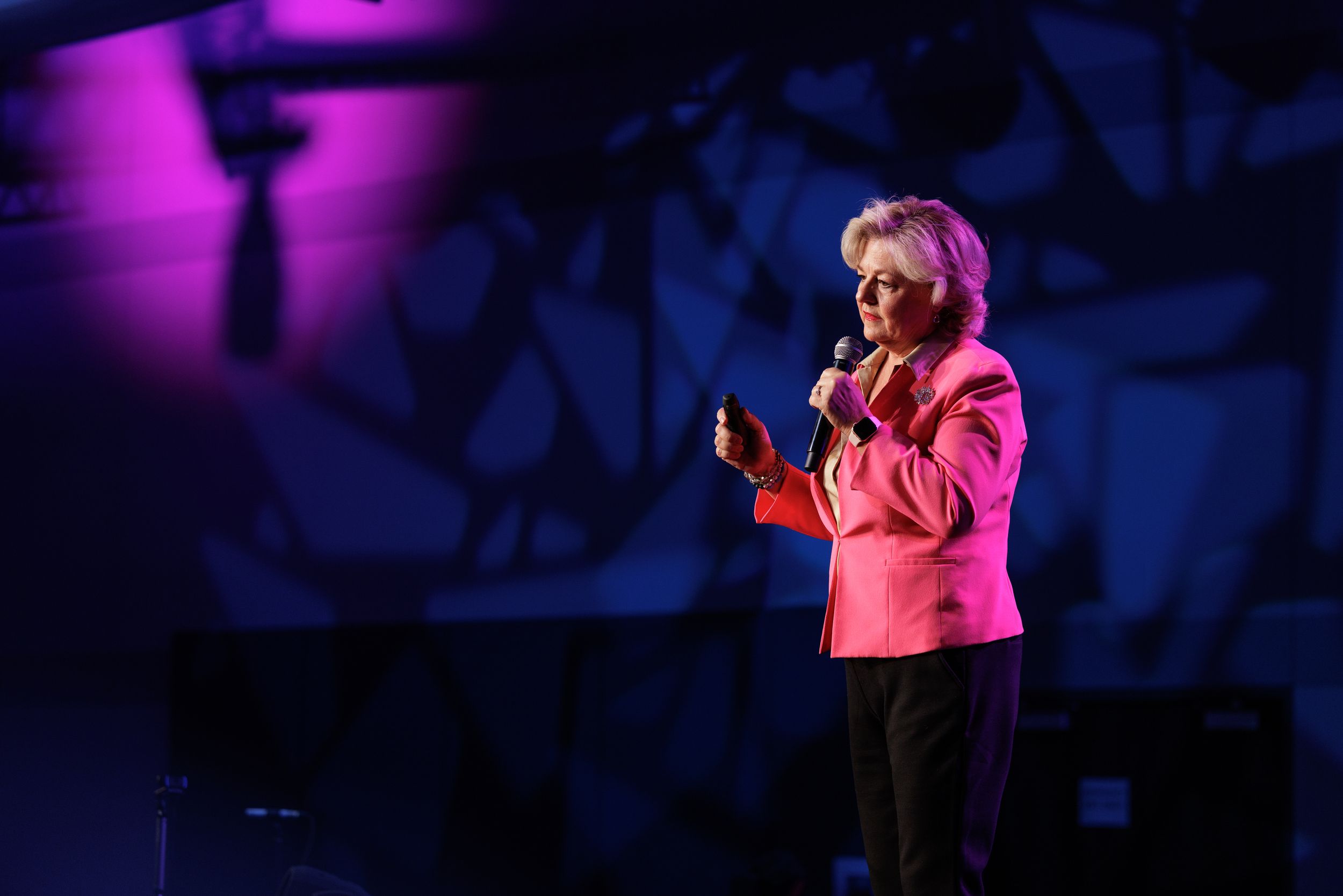 A speaker in a bright pink blazer gestures while giving a presentation on a darkened stage with purple lighting effects.