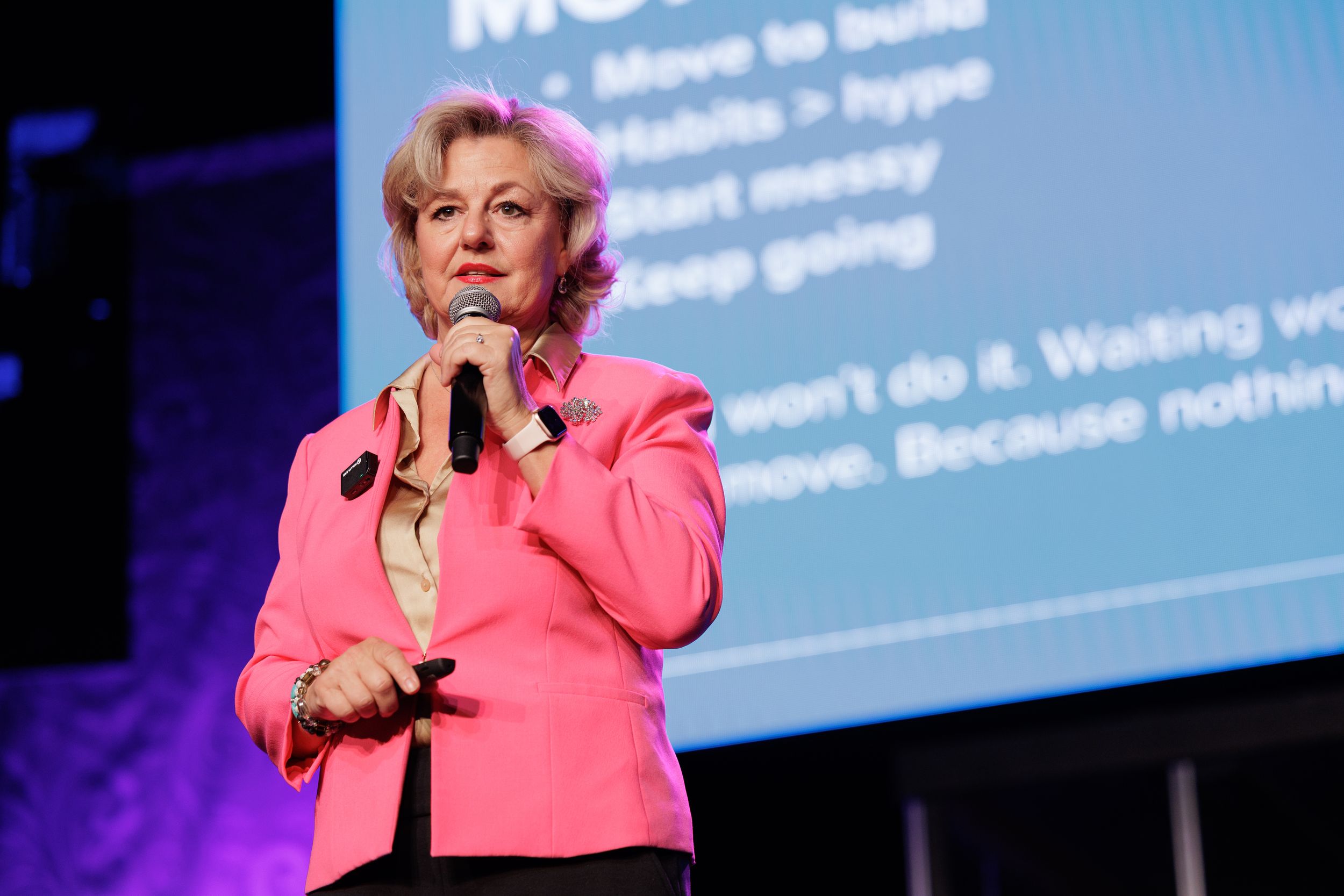 Professional speaker presenting on stage in bright pink blazer with large projection screen in background.