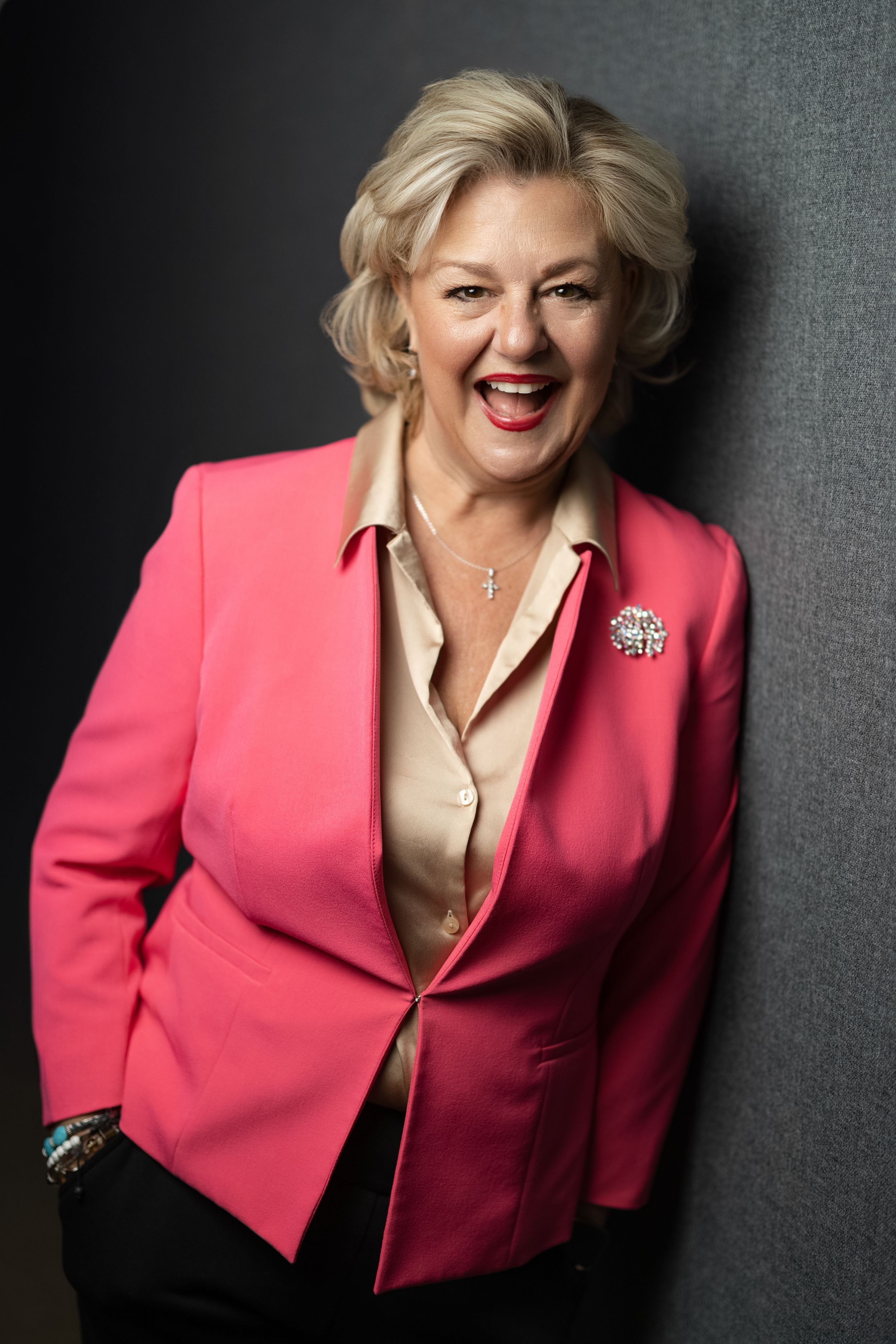 Professional portrait of a speaker wearing a vibrant pink blazer and beige blouse against a dark background.