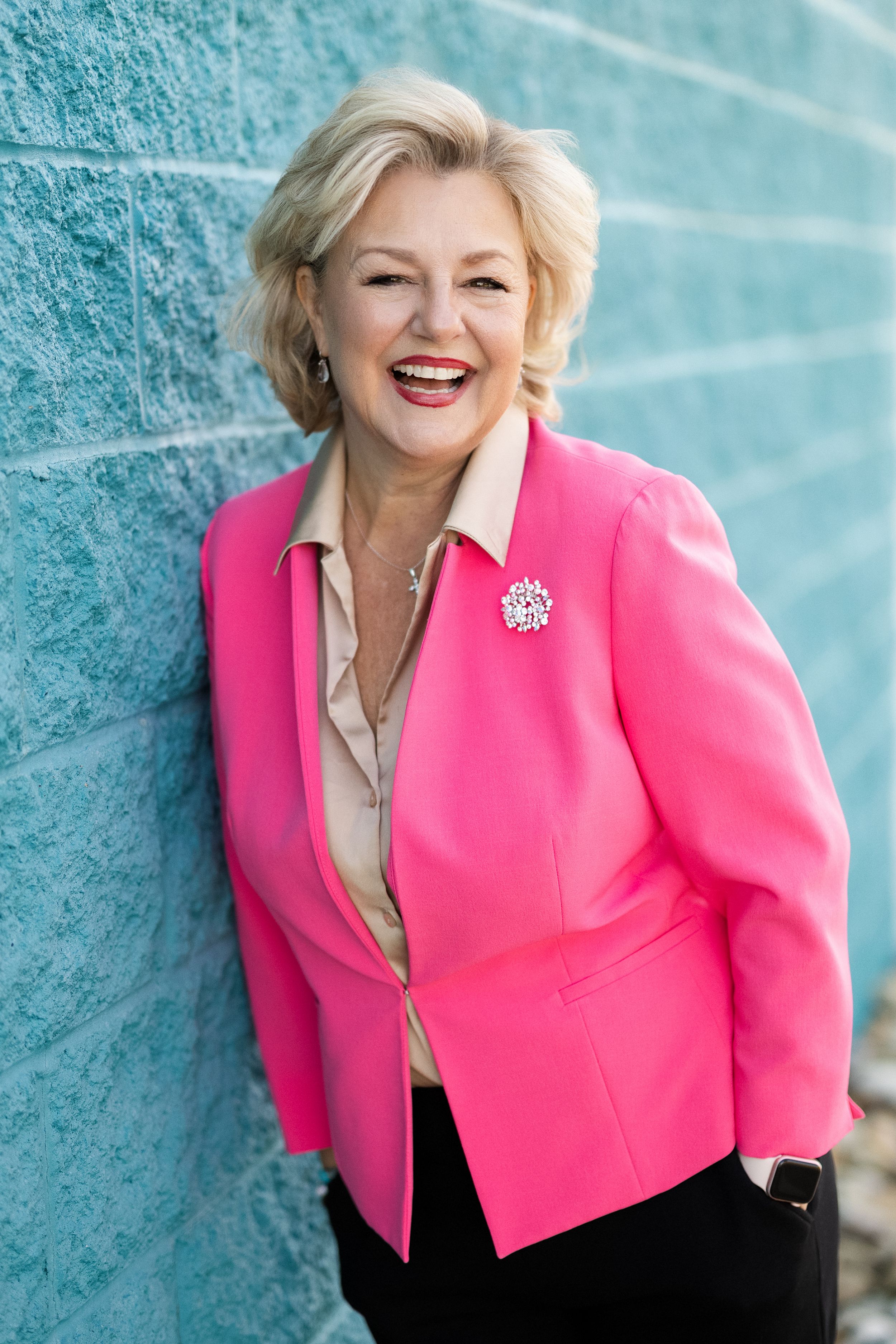 Blonde woman in vibrant pink blazer smiling against blue wall background with decorative brooch pin.