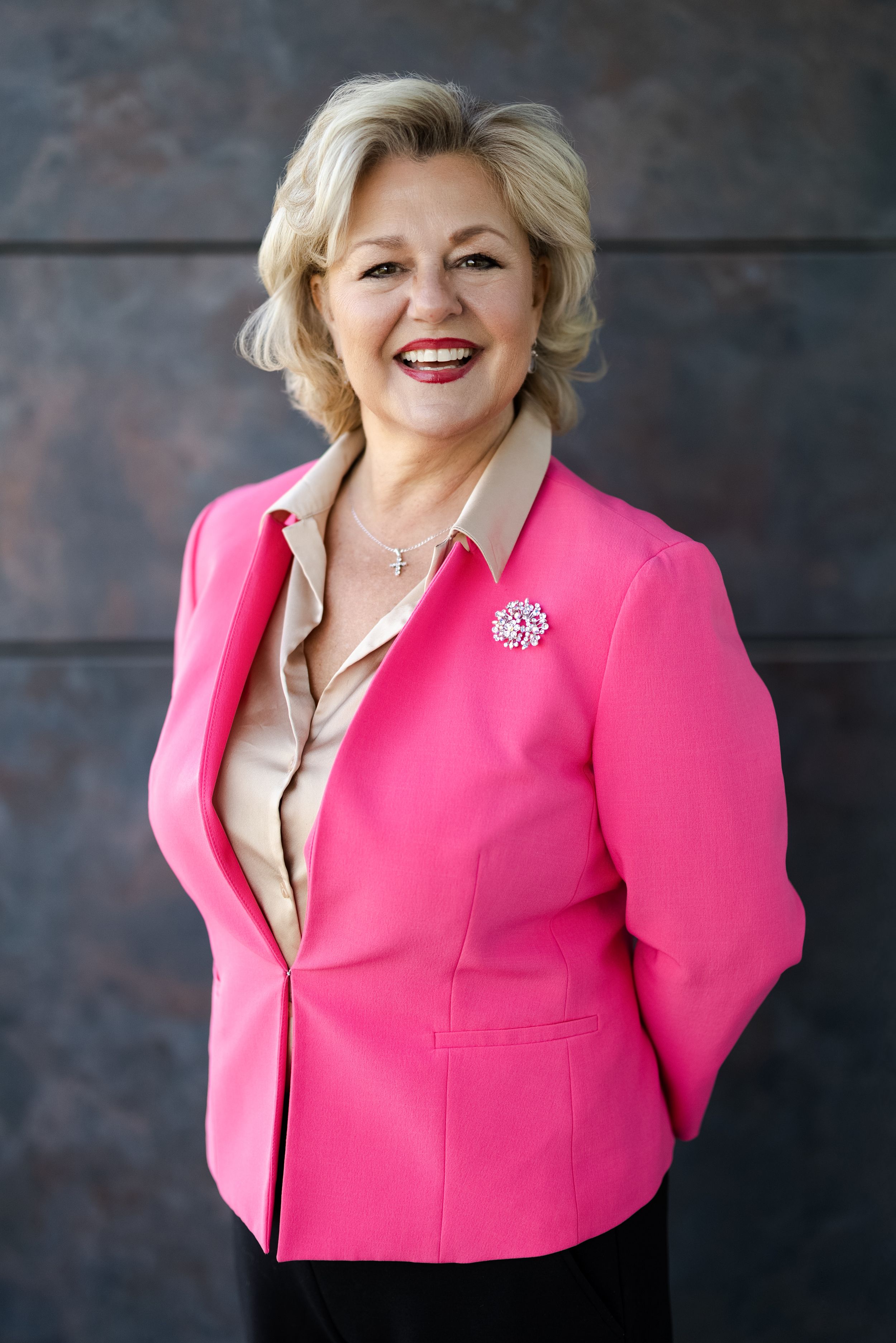 Professional portrait featuring bright pink blazer with decorative brooch pin and beige blouse against dark background.