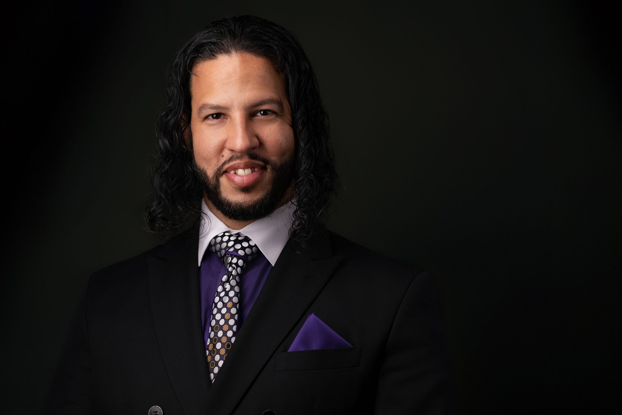 Professional headshot of a speaker wearing a dark suit with purple tie against a black backdrop.