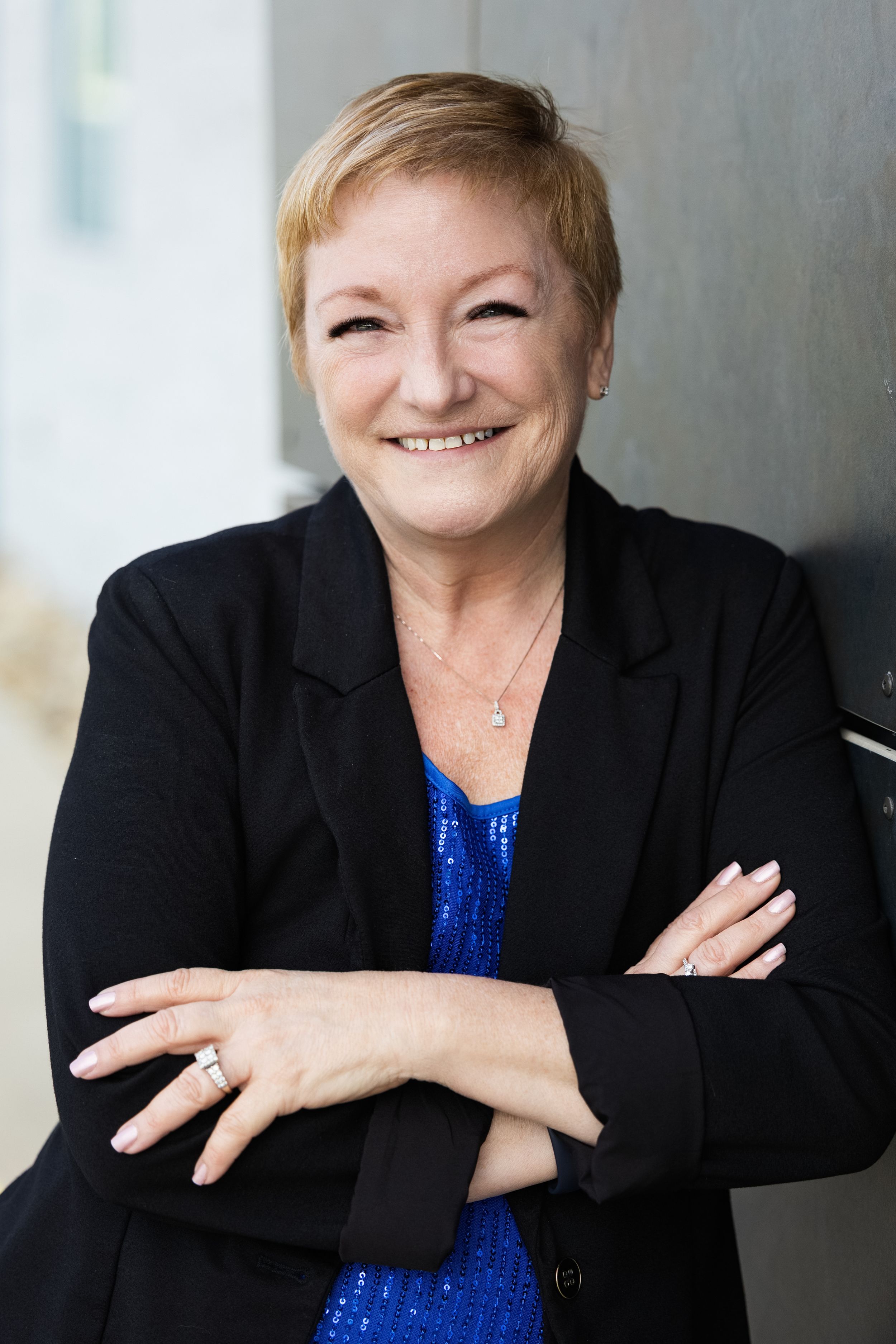 Professional business portrait of a person in a black blazer and blue top smiling warmly against a light background.