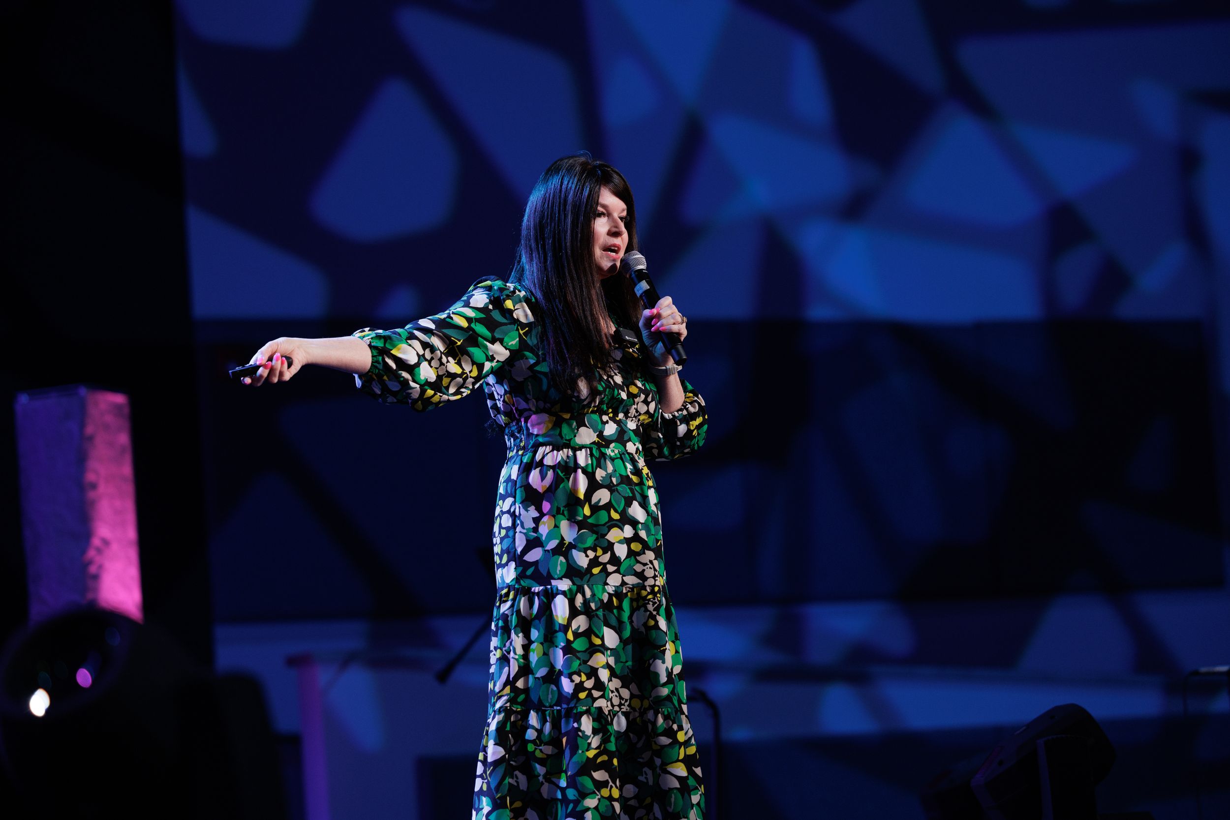 A performer in a long floral dress stands with outstretched arms on stage against a blue geometric backdrop.