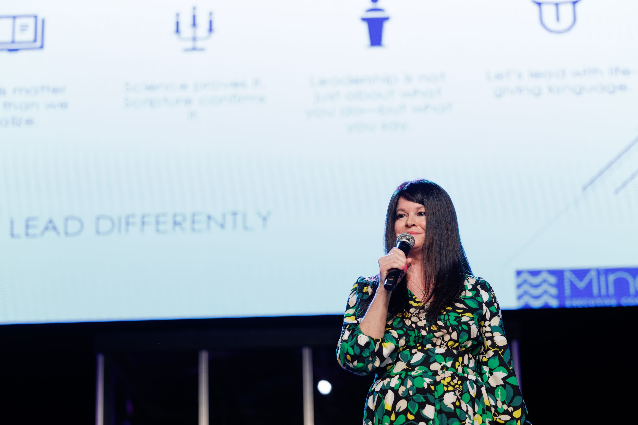 A speaker in a floral green dress addresses an audience from stage with presentation screens visible behind them.