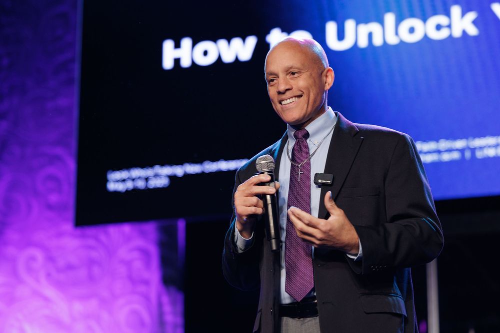 A speaker holds a microphone while presenting on stage with a blue illuminated backdrop and text display.