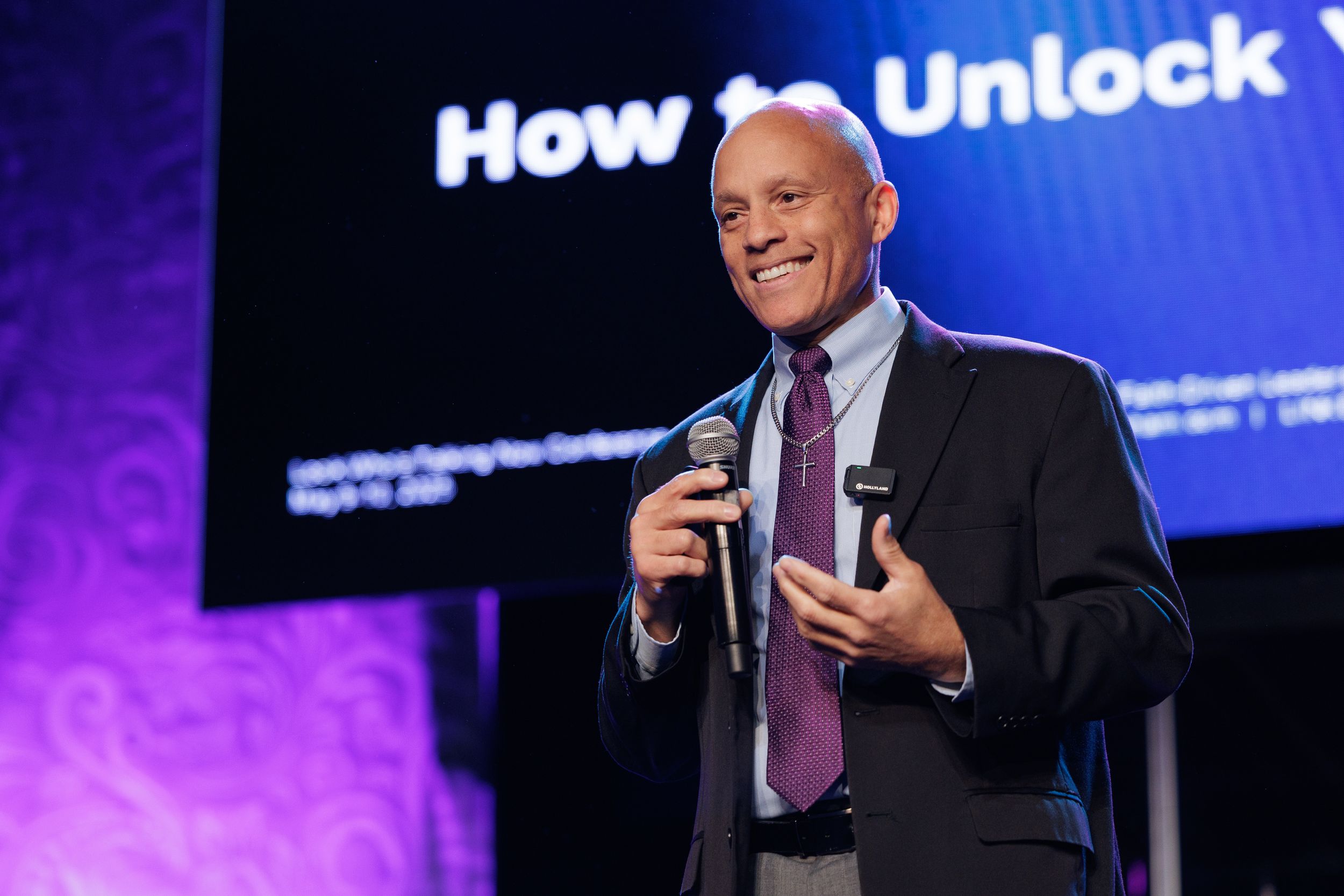 A speaker holds a microphone while presenting on stage with a blue illuminated backdrop and text display.