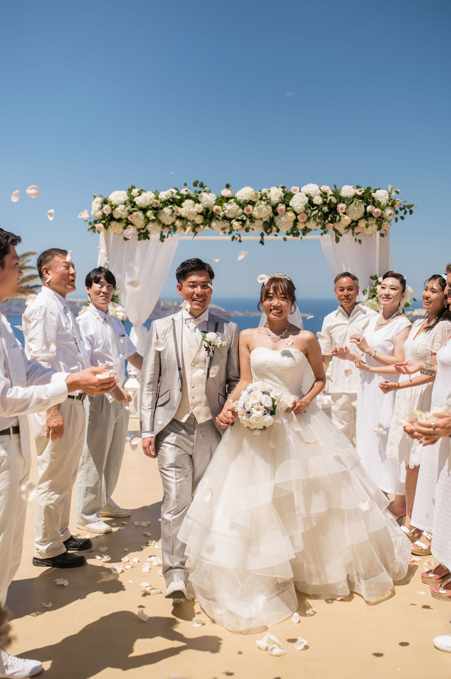 A beachfront wedding ceremony with the happy couple and wedding party in white attire standing under a floral arch.