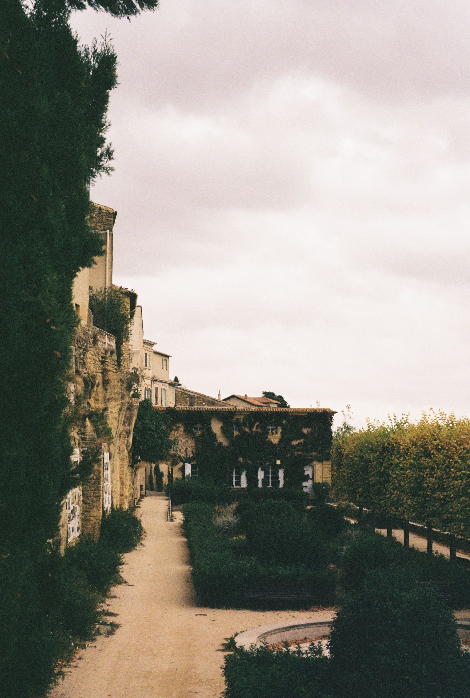 Historic garden pathway lined with trees and stone architecture in muted tones.