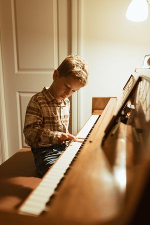 A person plays piano in warm lighting while sitting at a wooden upright piano in a cozy home setting.