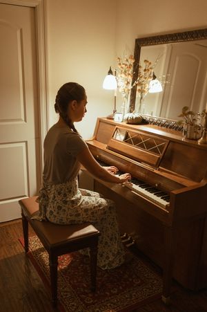 A pianist in a floral dress plays an upright piano in a warmly lit room with ornate decor and an oriental rug.