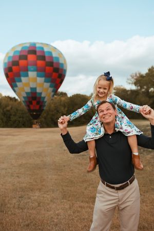 Hot air balloons float in background as people play and pose in a grassy field on sunny autumn day.
