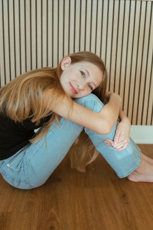 Portrait of someone in light blue jeans and black top sitting casually against wooden paneled wall.